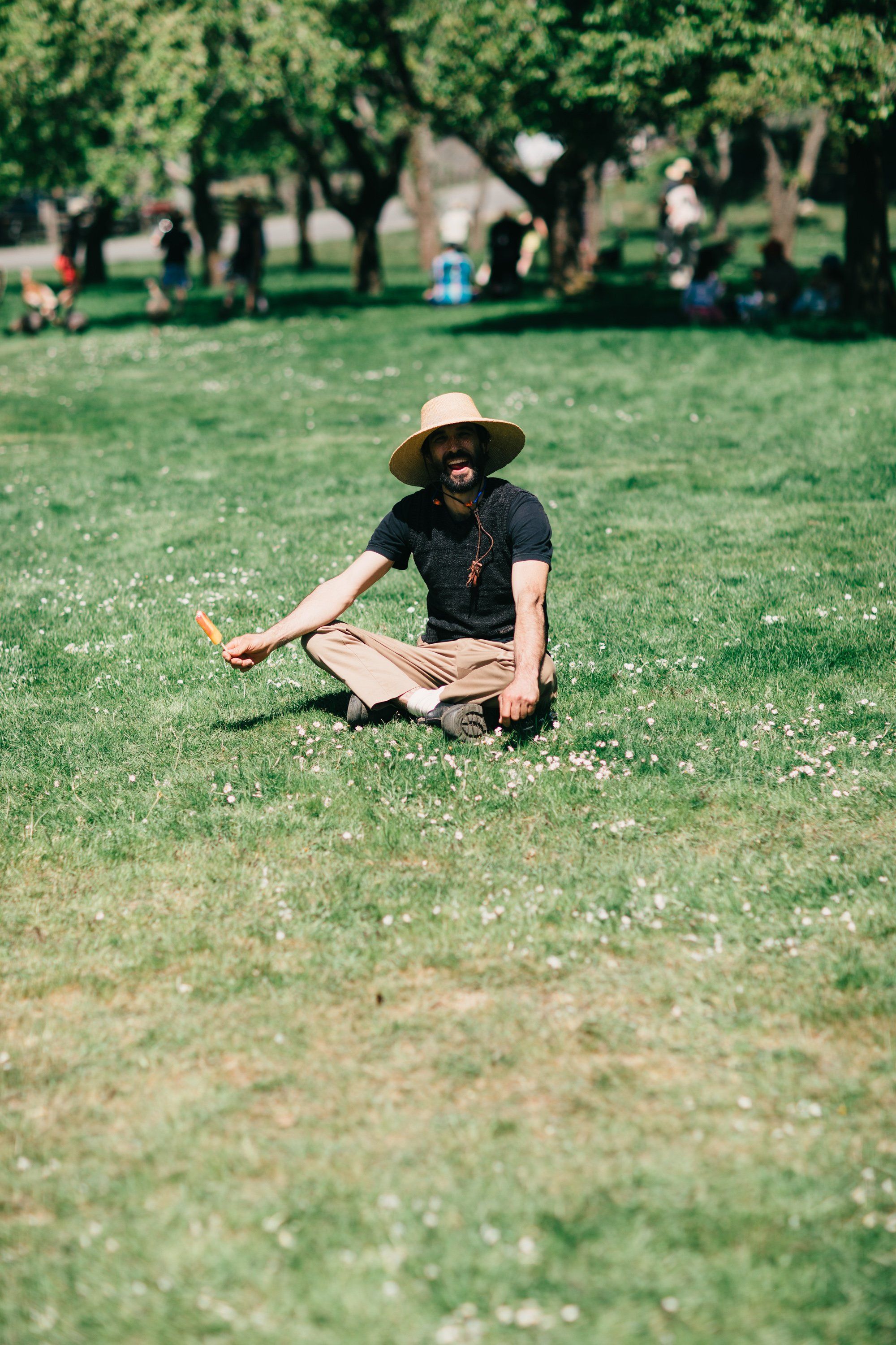 Person in black shirt and straw hat sits cross-legged on sunny grass, holding orange produce at Ruckle Farm