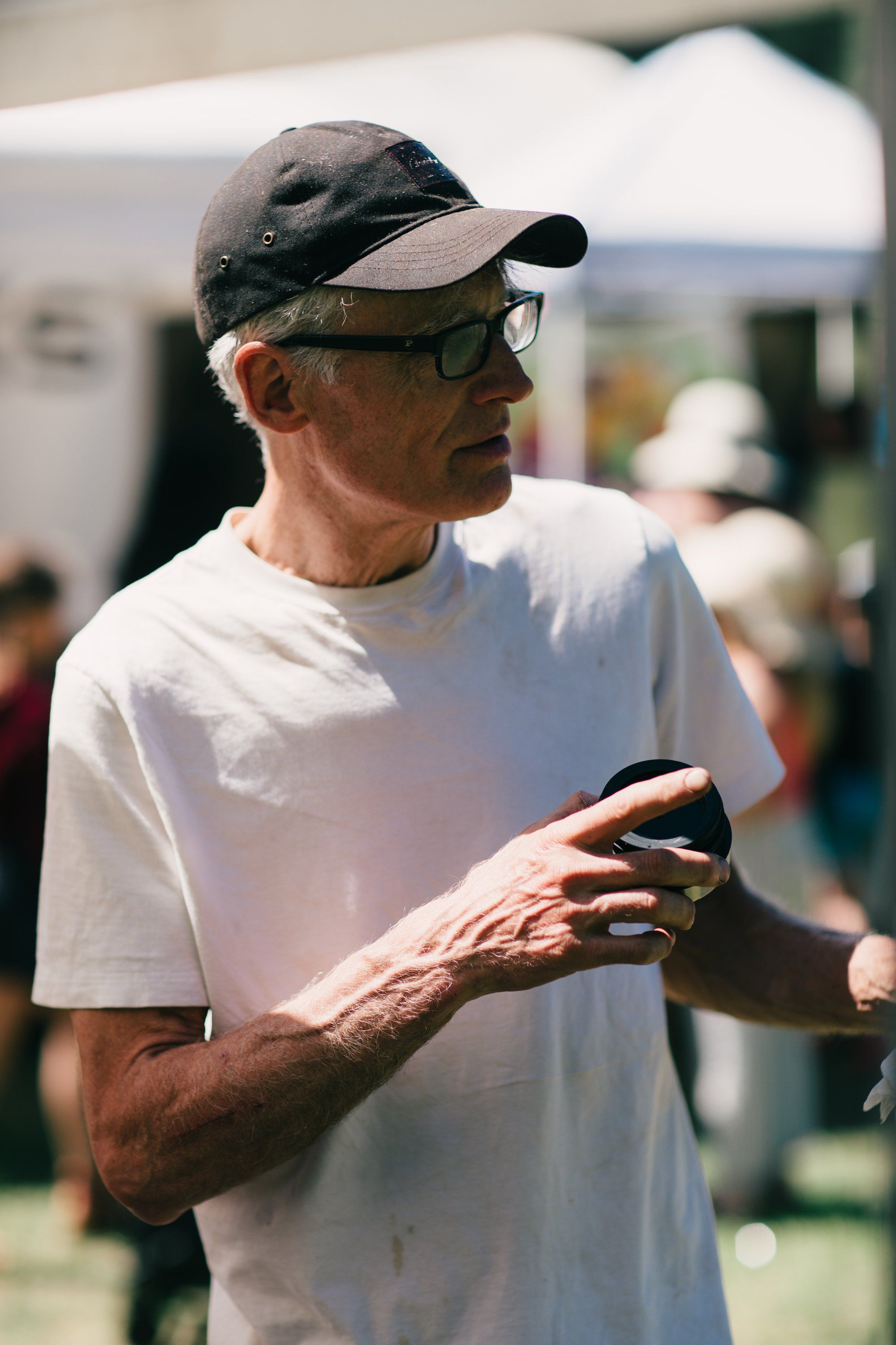 Older man in white t-shirt and black cap holding phone at outdoor farm event, golden hour light