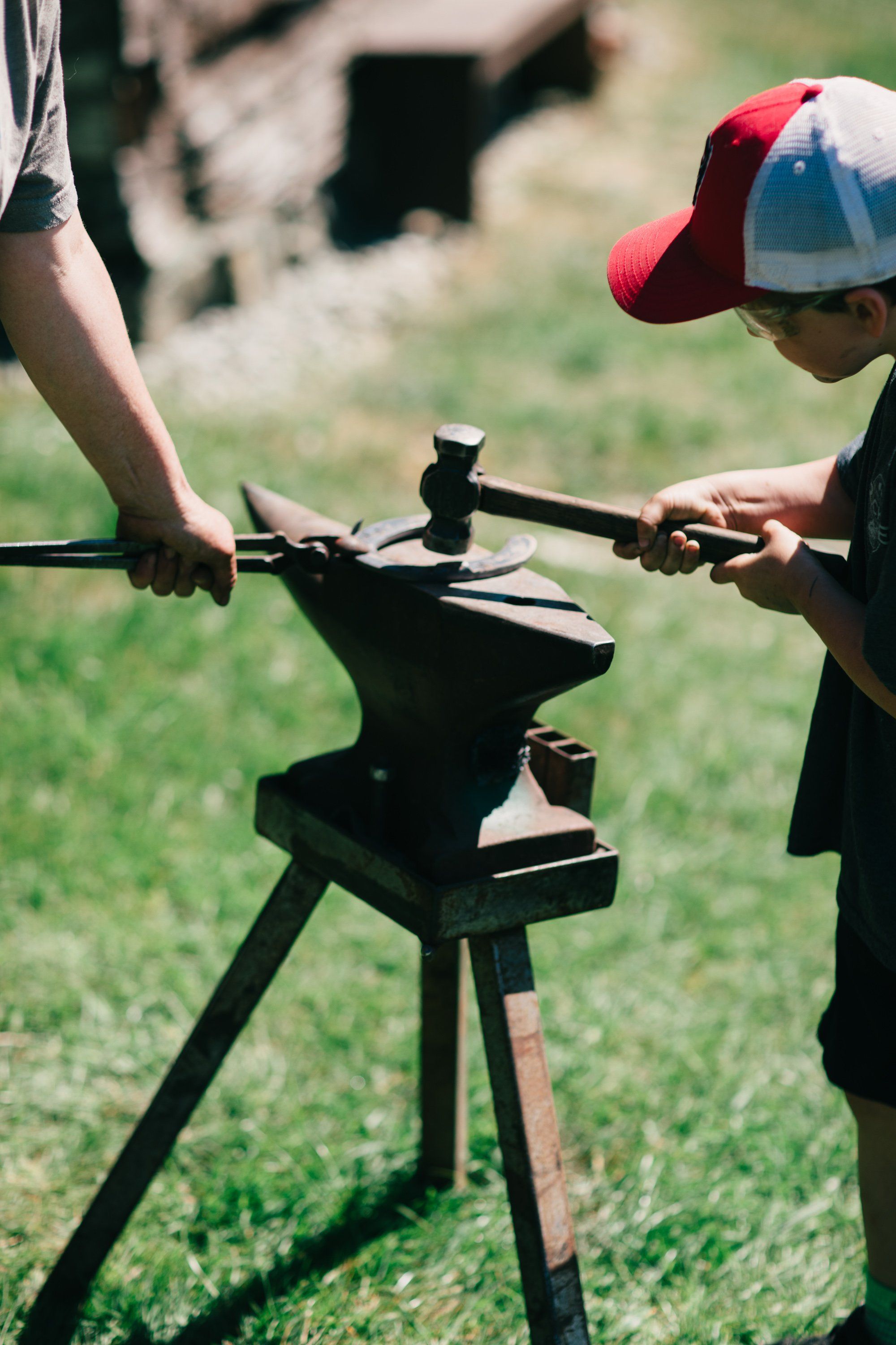 Child in red cap learning to use a blacksmith's vise on grass, guided by an adult's hands