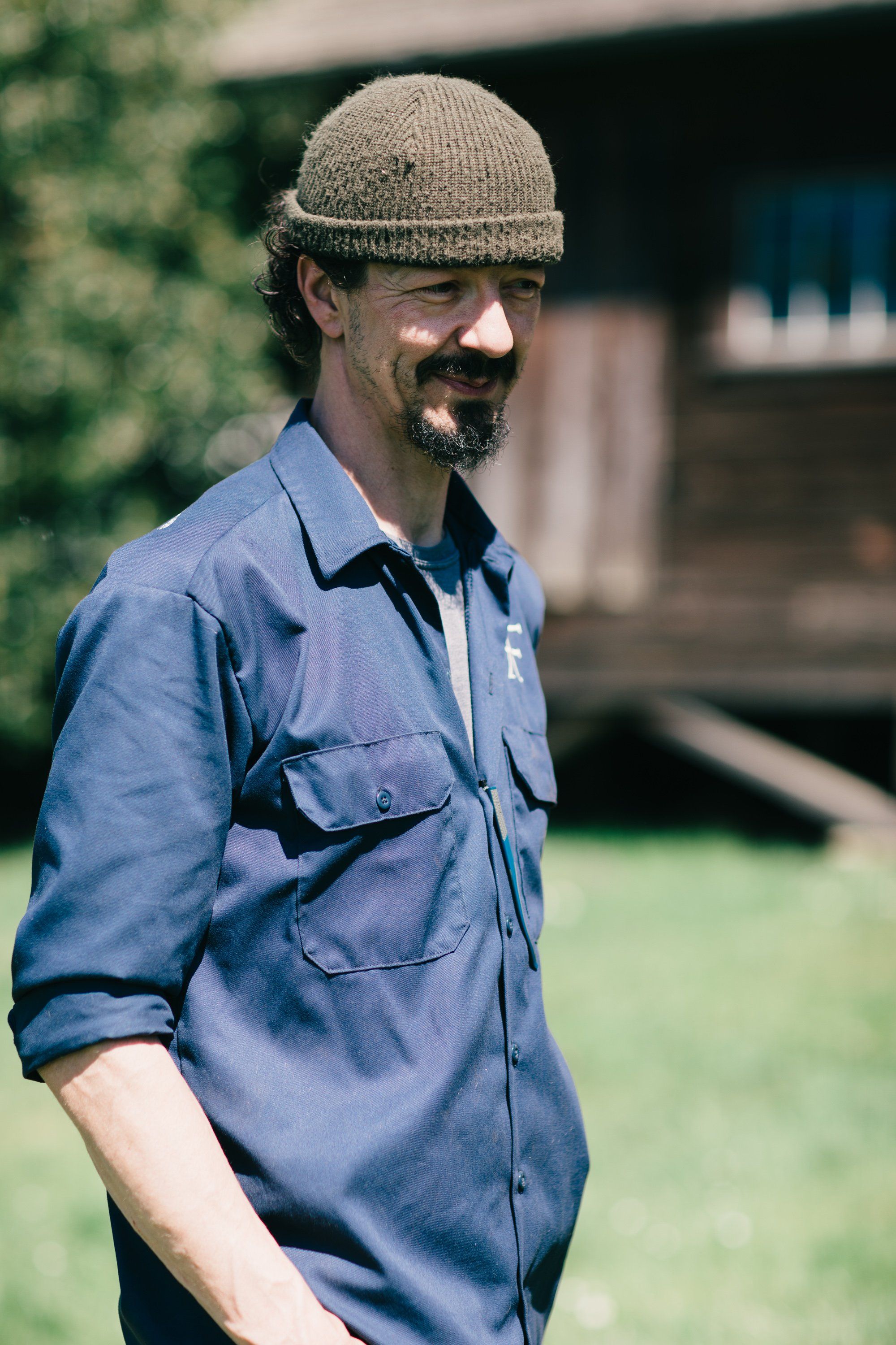 Man in blue work shirt and knit beanie smiling at camera in sunlit farm setting with wooden buildings behind
