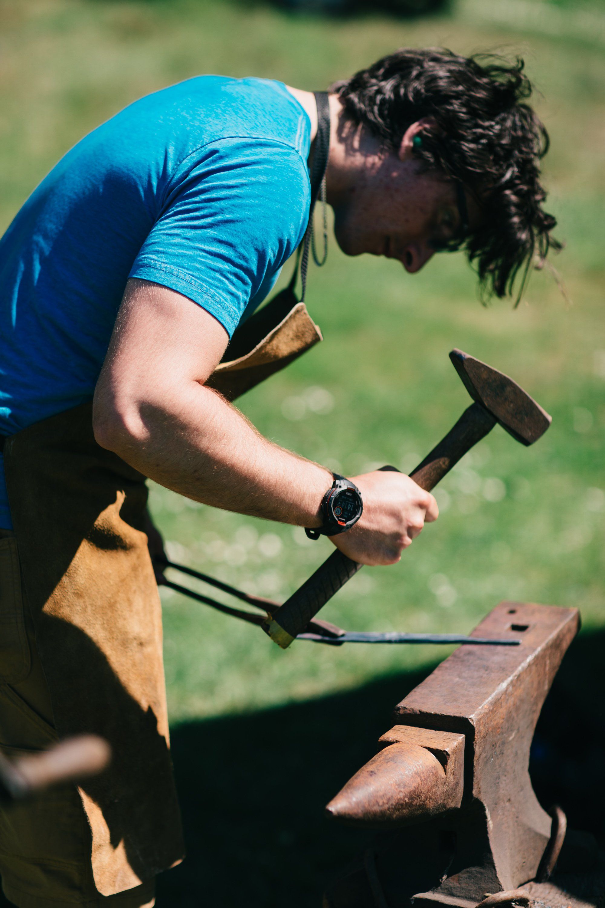 Man in blue shirt hammering at an anvil with focused intensity, golden afternoon light illuminating the rural farm setting