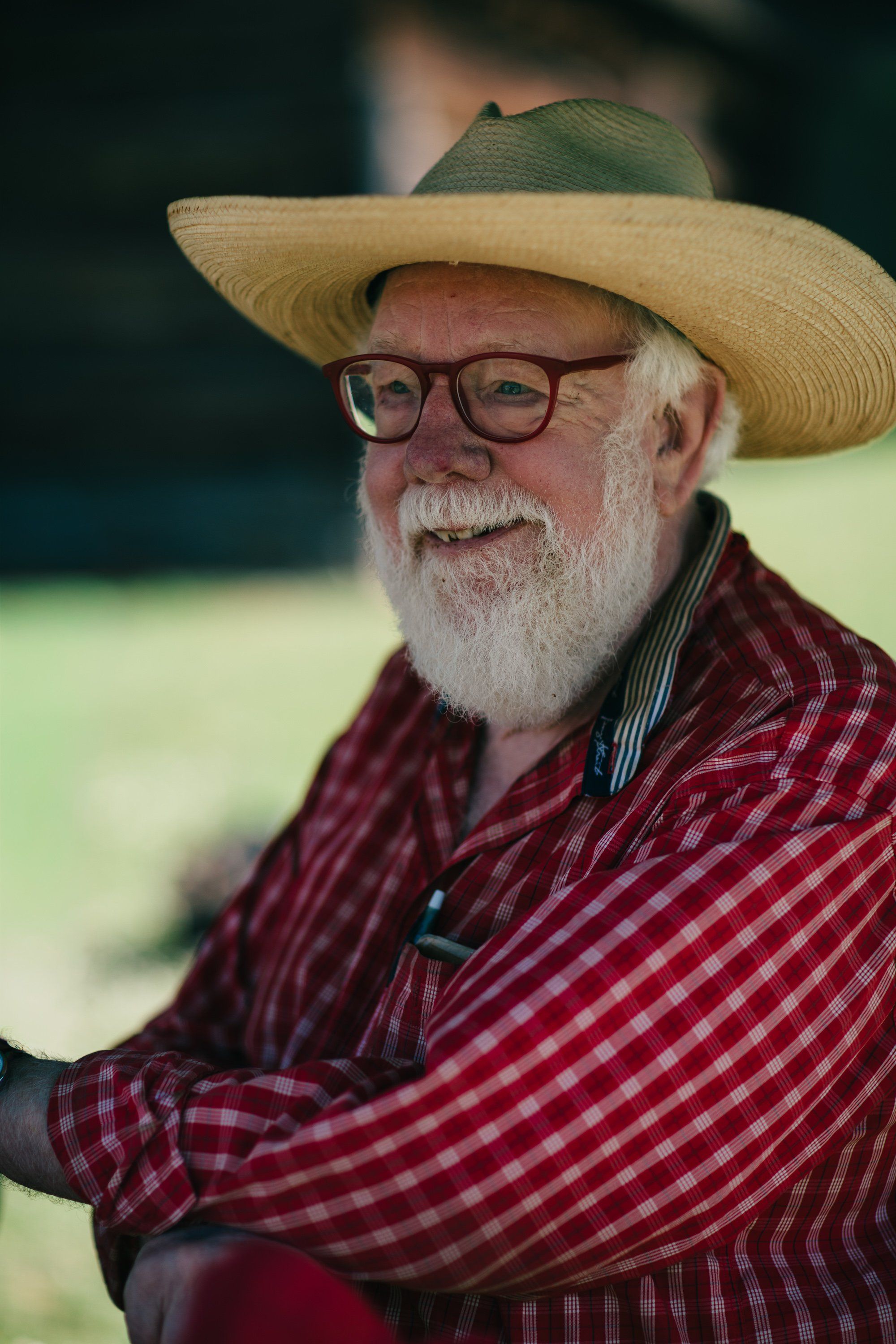 Bearded farmer in wire glasses and wide-brimmed straw hat smiles warmly while holding a farming tool, wearing a burgundy plaid shirt against a soft-focus gre…