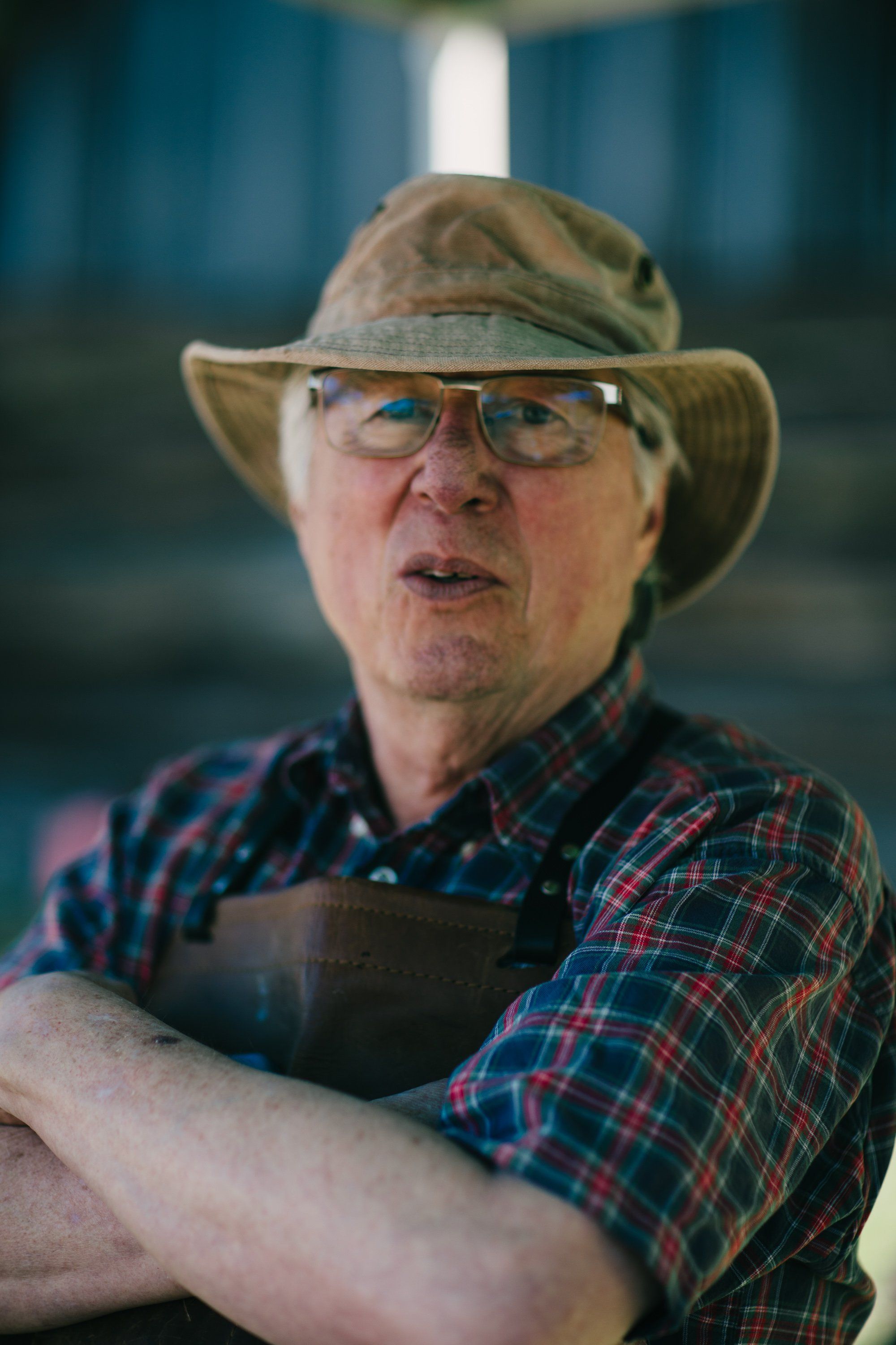 Farmer in plaid shirt and canvas hat with glasses stands in barn doorway, arms crossed, morning light illuminating weathered face and calm expression