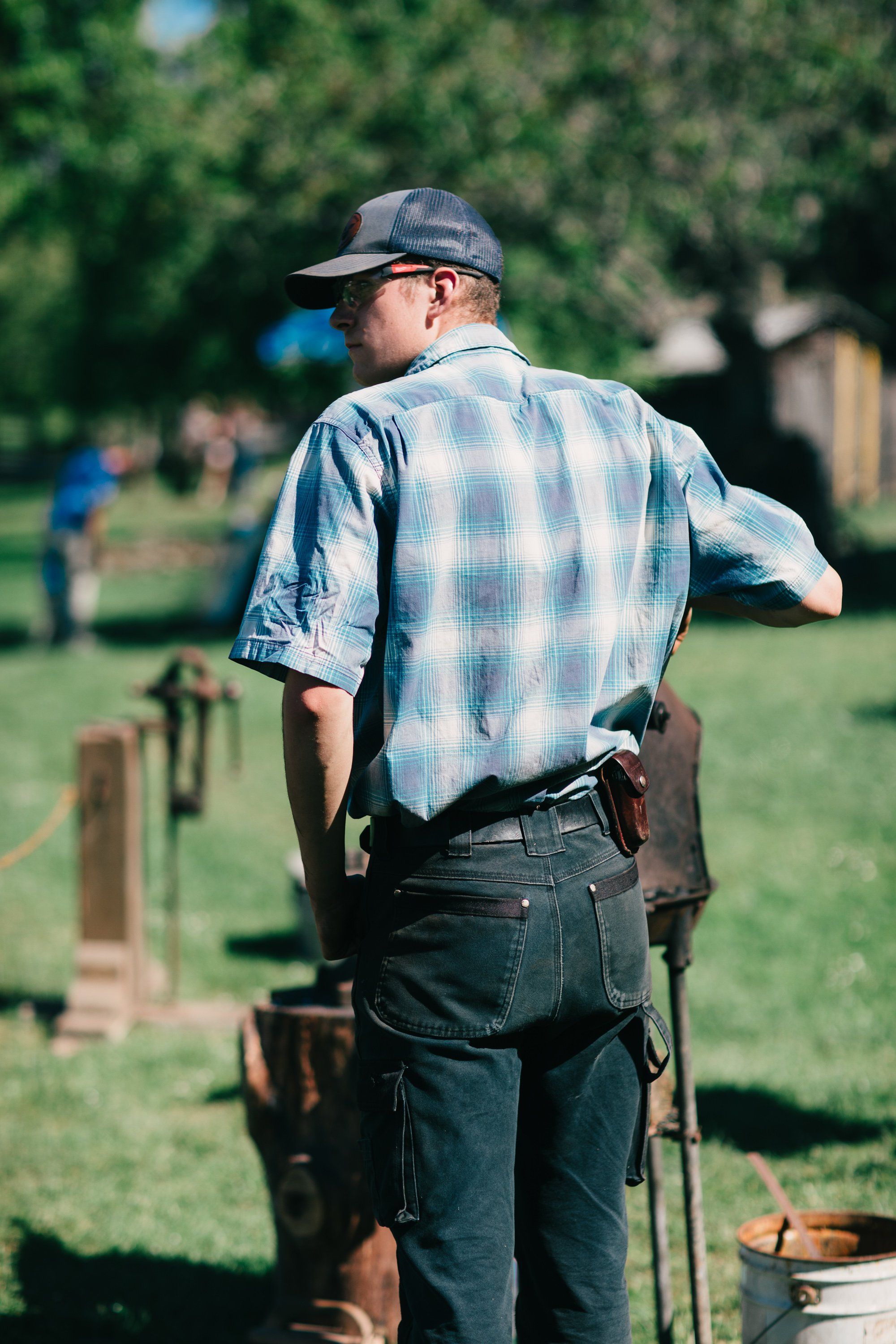 Farmer in blue plaid shirt walks across sunny field at Ruckle Farm, tool in hand, with blurred trees and structures behind