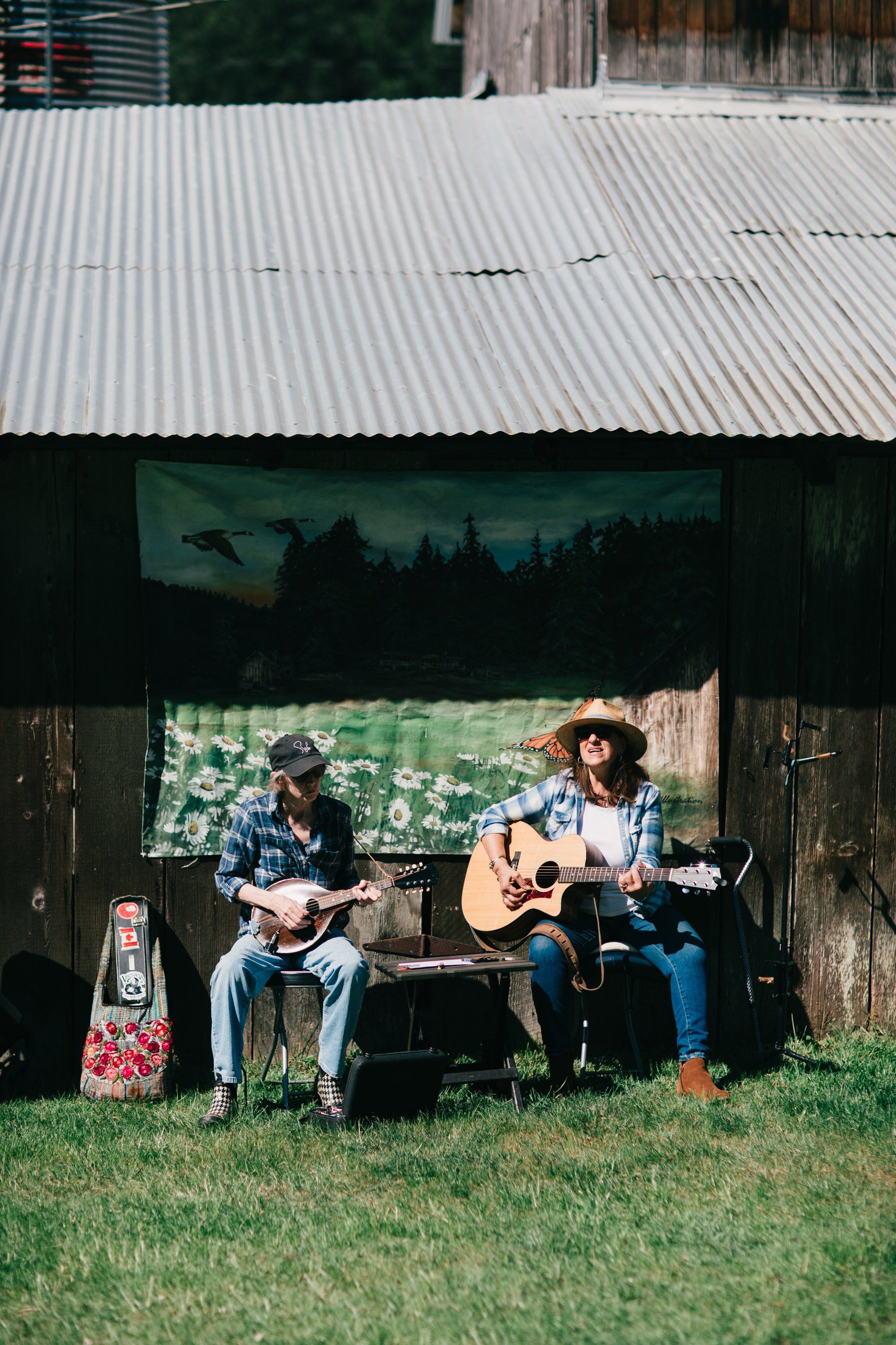 Two musicians with guitars perform in golden afternoon light beneath a corrugated metal barn roof, seated on grass with scenic backdrop
