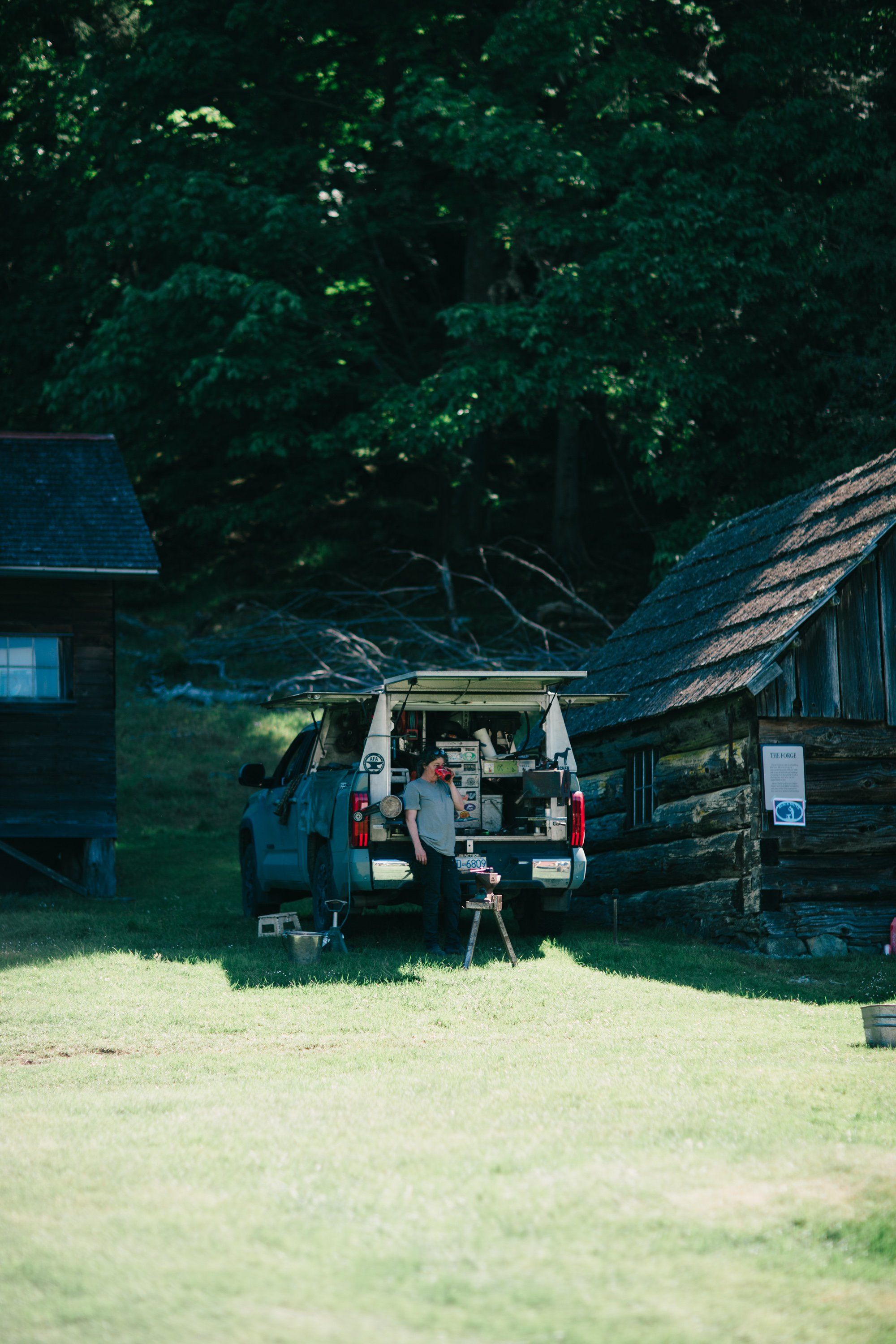Vintage van parked between rustic farm buildings with dense green forest canopy, dappled sunlight across sandy ground