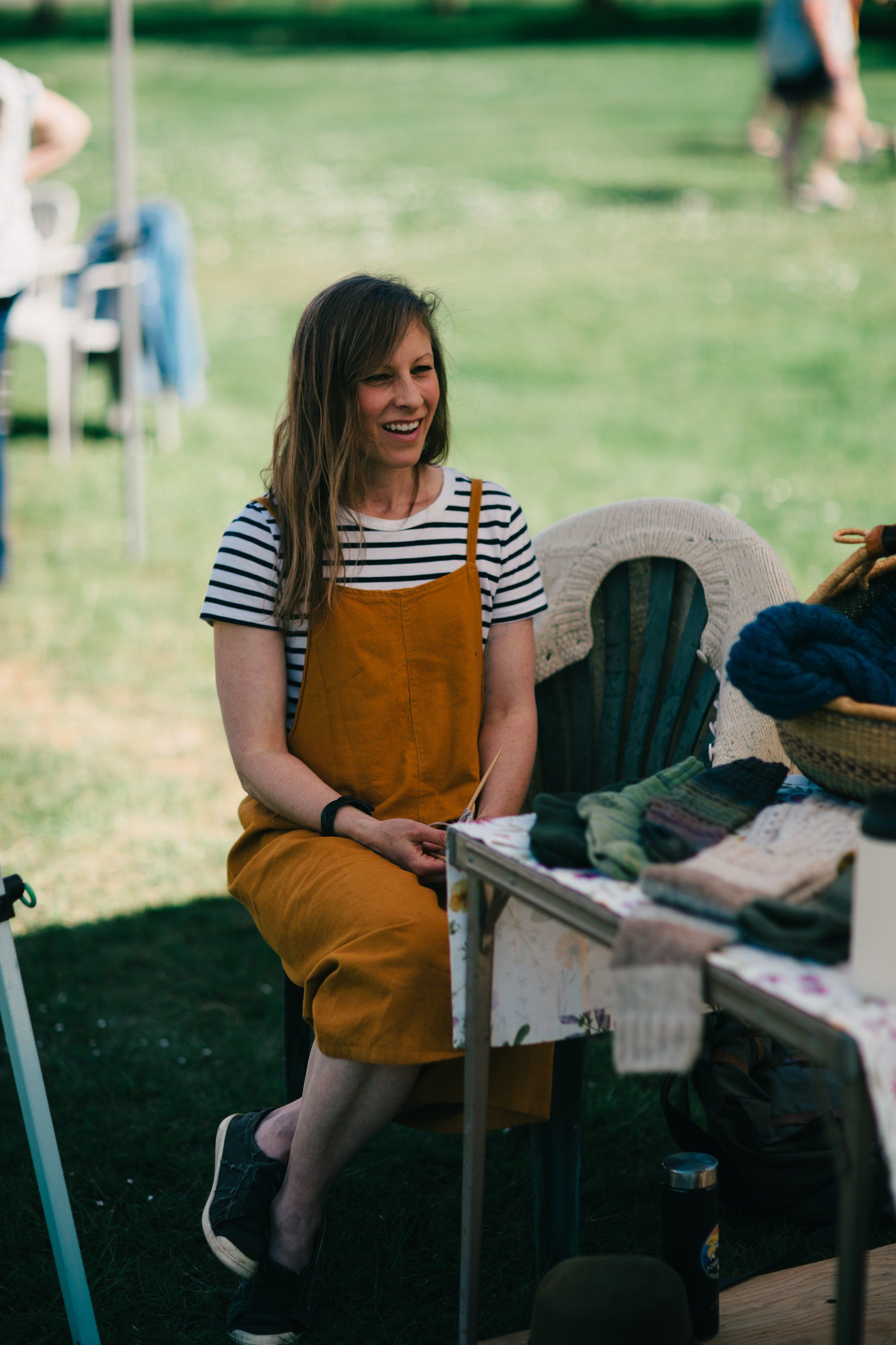 Woman in mustard overalls and striped shirt sits at a wooden table on a sunny farm, smiling while holding a tool, with green fields and other people visible…