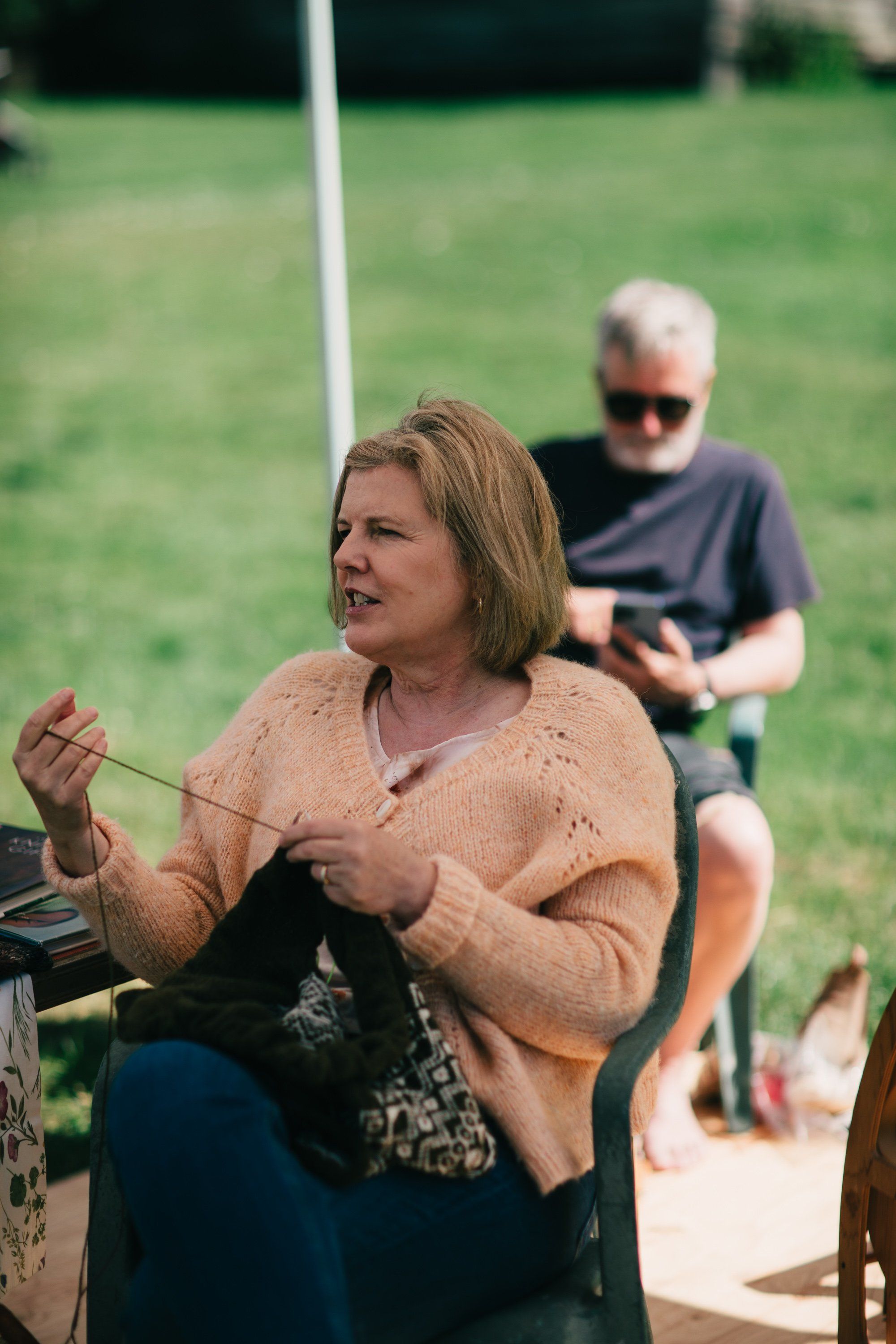 Woman in tan cardigan gestures while speaking at Ruckle Farm, with man in sunglasses and spectators in soft afternoon light
