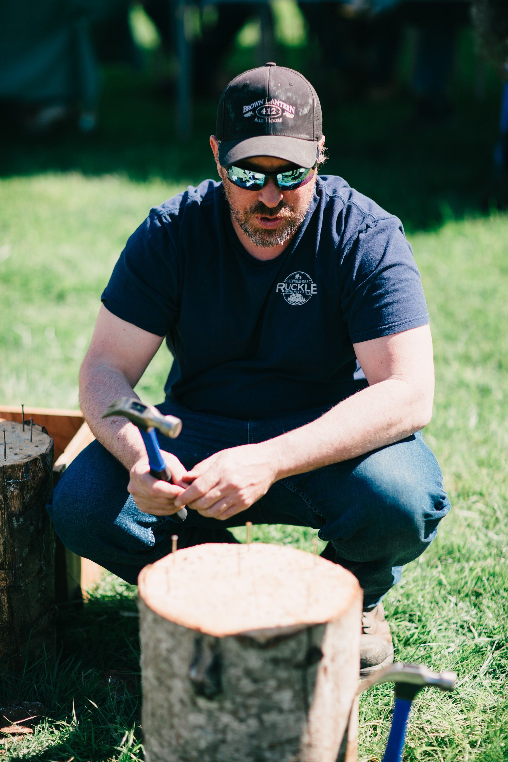 Man in dark shirt and cap carving wood with knife at rustic log table in sunlit grass field