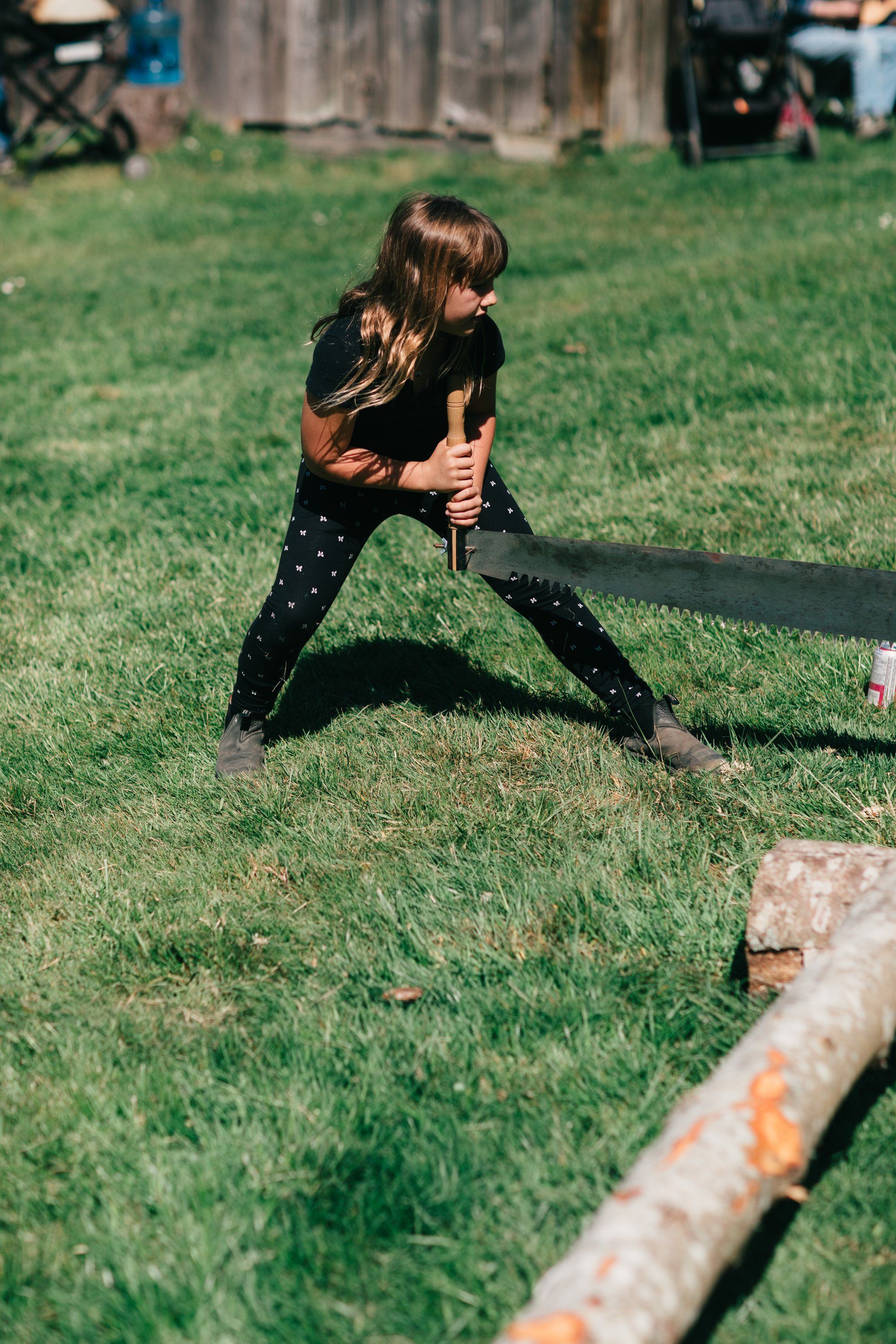 Child in dark shirt and patterned leggings lunges toward a wooden log on sunny grass at Ruckle Farm
