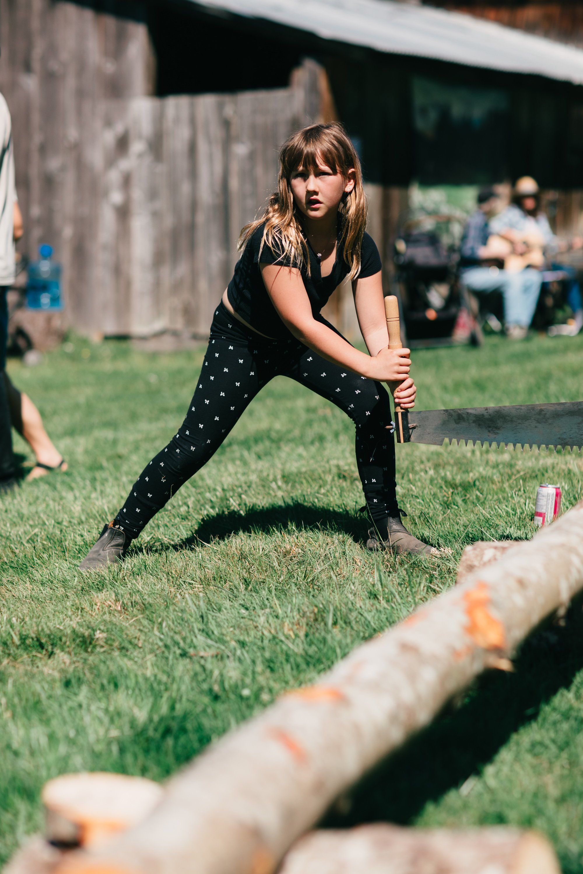 Woman in black polka-dot pants stretches toward a log during outdoor farm games, backlit by afternoon sun