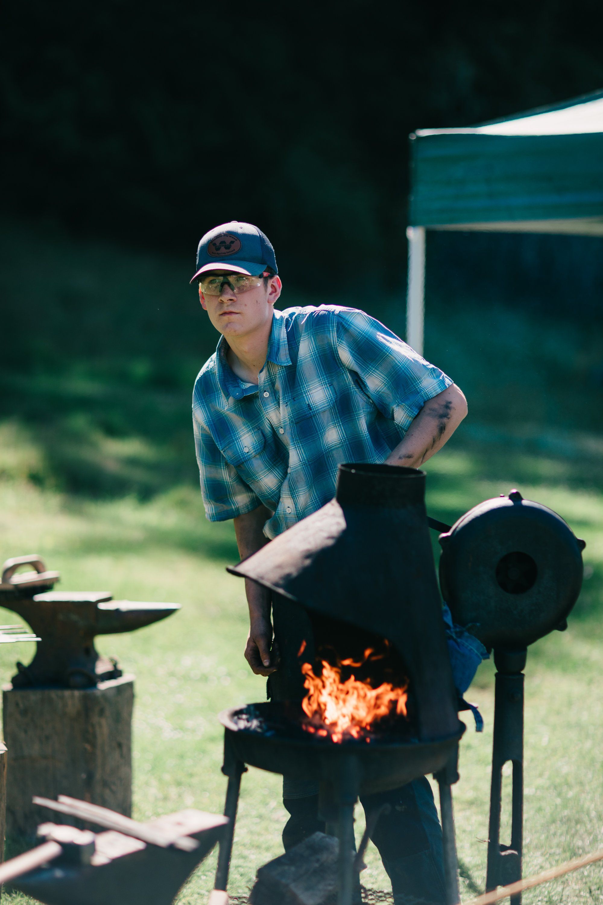 Man in cap and plaid shirt tends a smoking barrel stove amid pastoral farmland and morning light