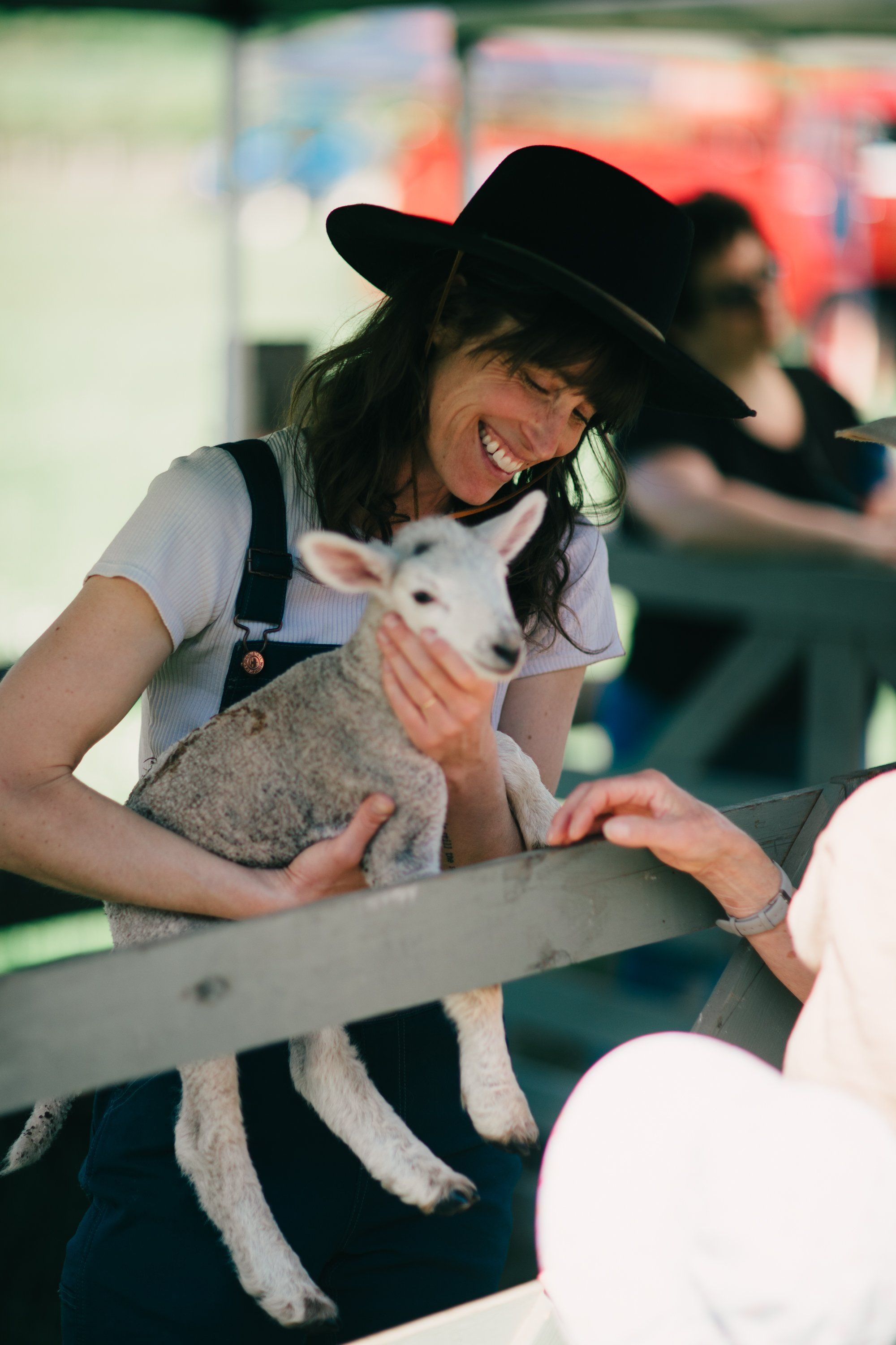 Woman in cowboy hat smiling while holding a young goat at a farm event, natural daylight