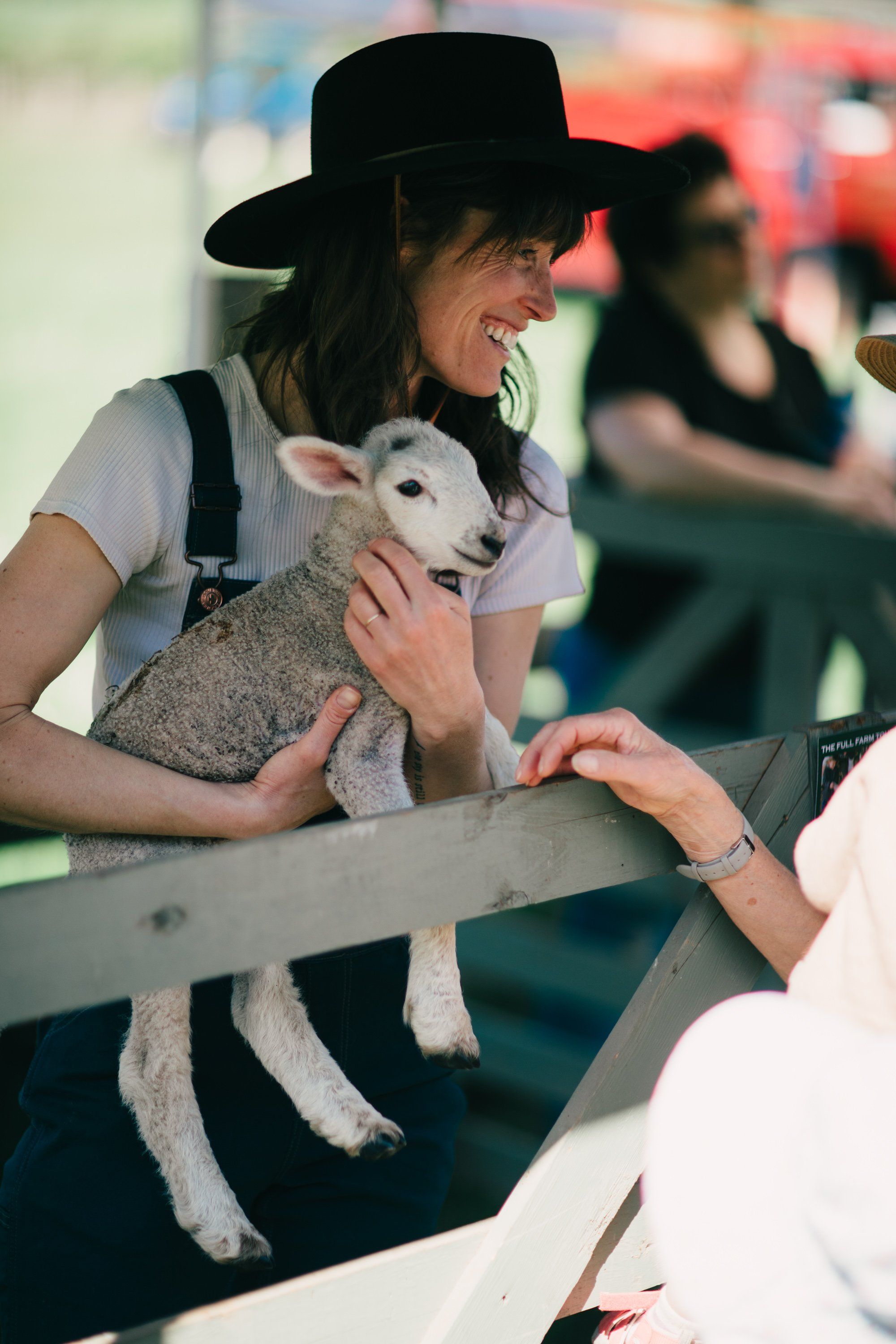 Woman in black hat holding young lamb at farm fence, smiling as visitor reaches toward the animal