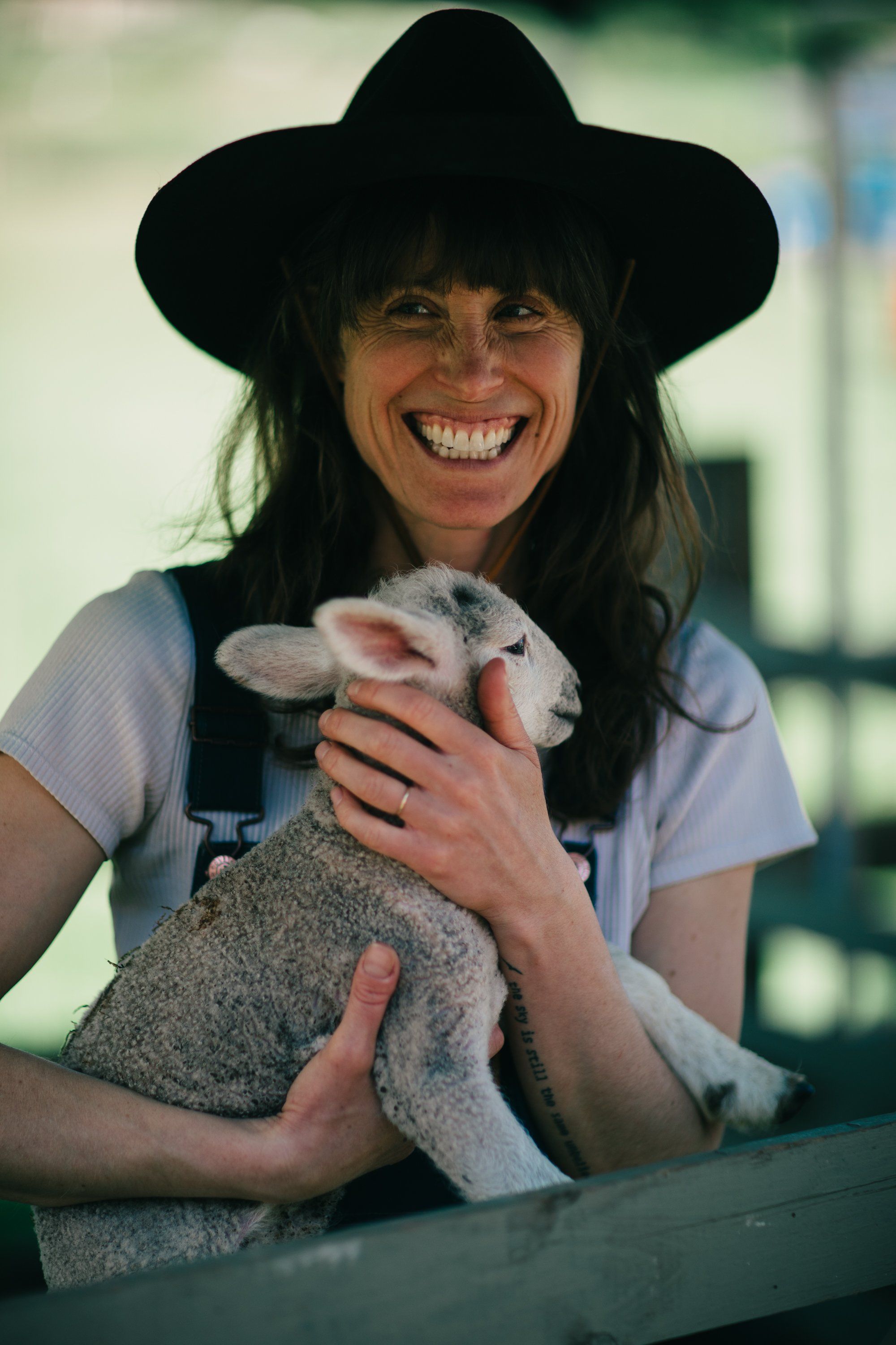 Woman in black cowboy hat holding young lamb, beaming at camera in bright farmyard light