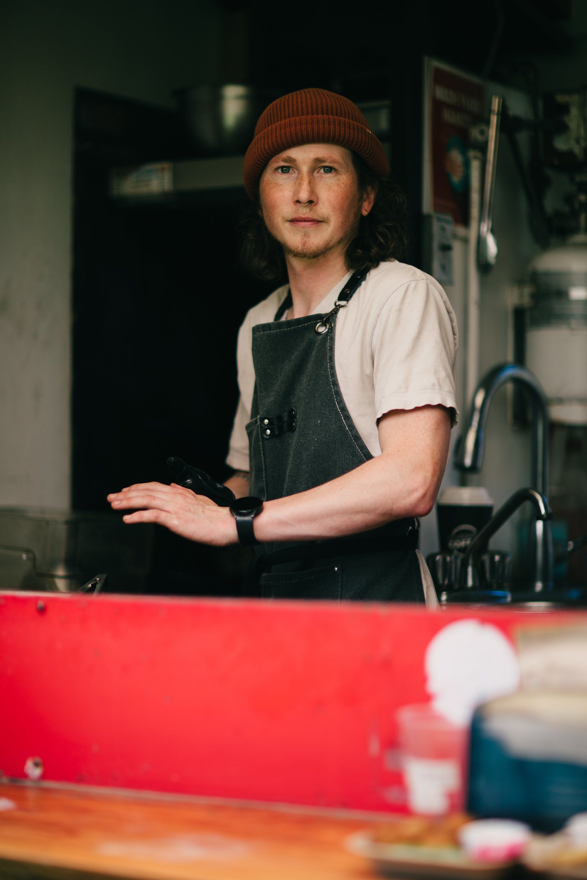 Man in rust beanie and denim apron stands in a farm workspace, gesturing while speaking, warm natural light illuminating his contemplative expression