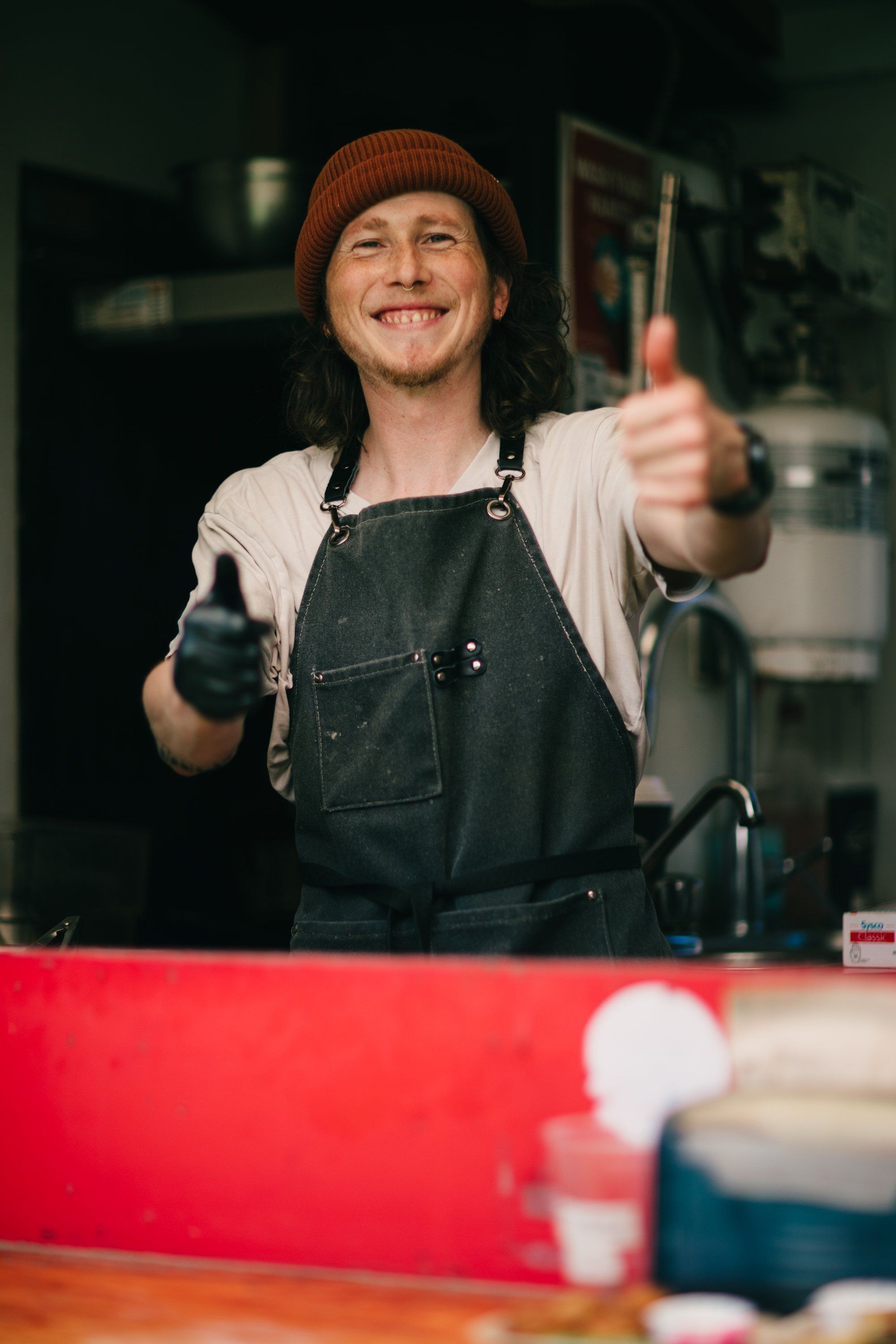 Person in denim apron and rust beanie gives double thumbs up at a wooden workbench in a rustic workshop setting