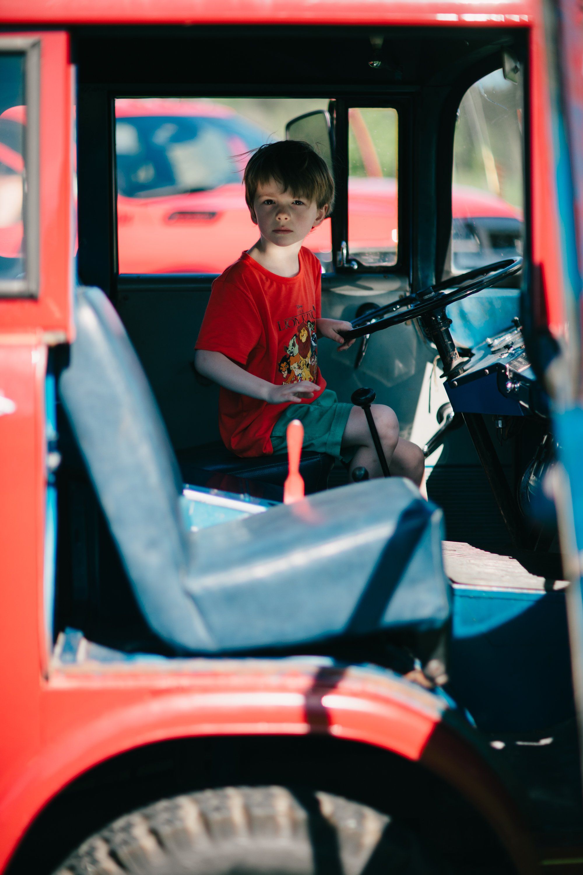 Young boy in red shirt sits in bright red vintage tractor cab, holding steering wheel with warm afternoon light filtering through windows