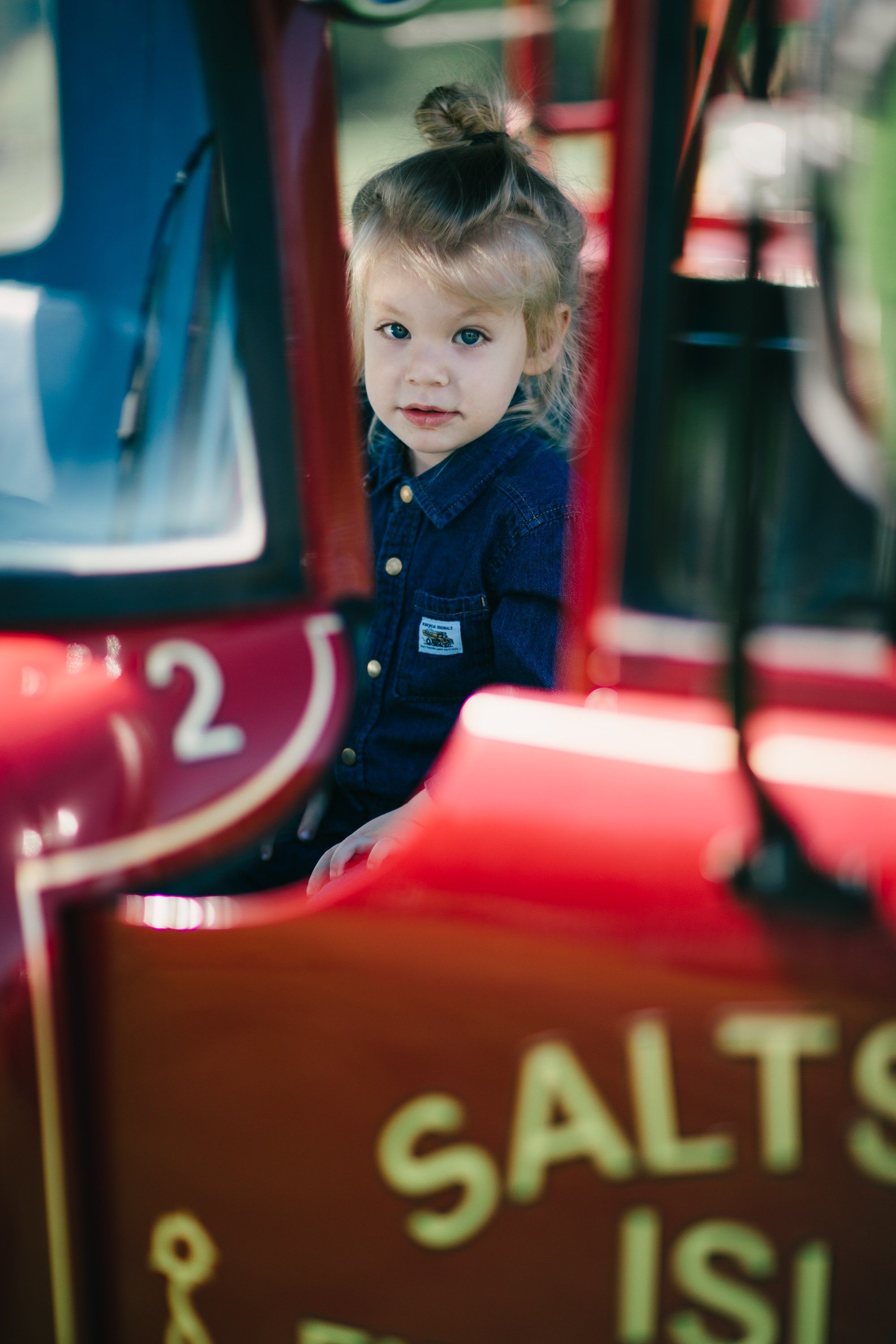 Toddler in navy shirt peeks through red farm equipment at Ruckle Farm, golden afternoon light illuminating her curious expression