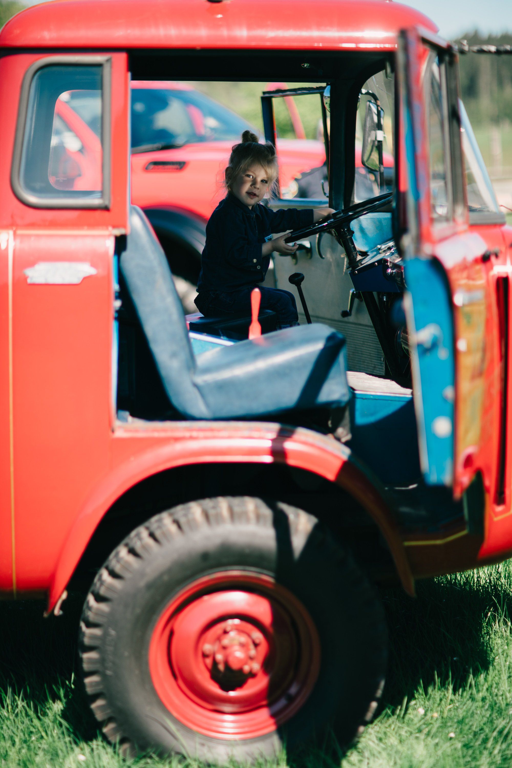 Child sits at the wheel of a vintage red farm truck, hands on steering wheel, morning light filtering through the cab window