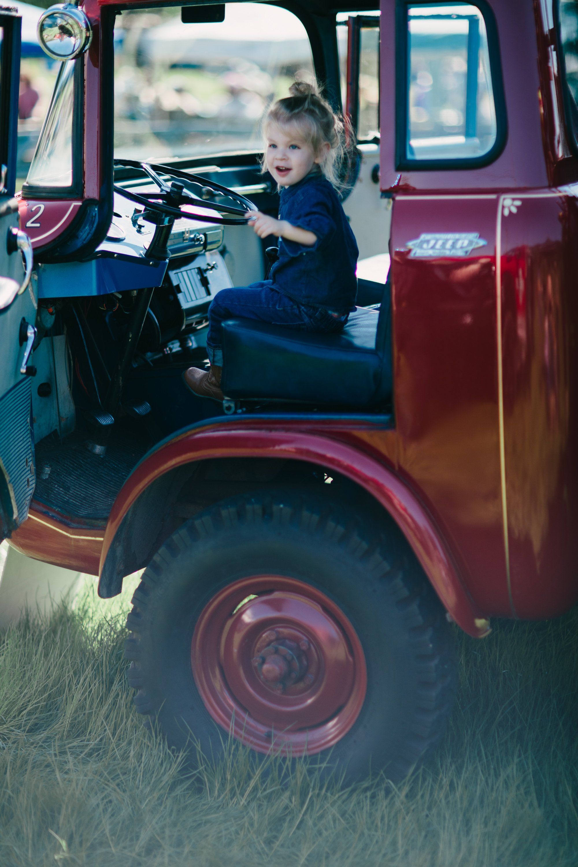 Young child at the helm of a weathered red farm truck, hands on steering wheel in golden afternoon light