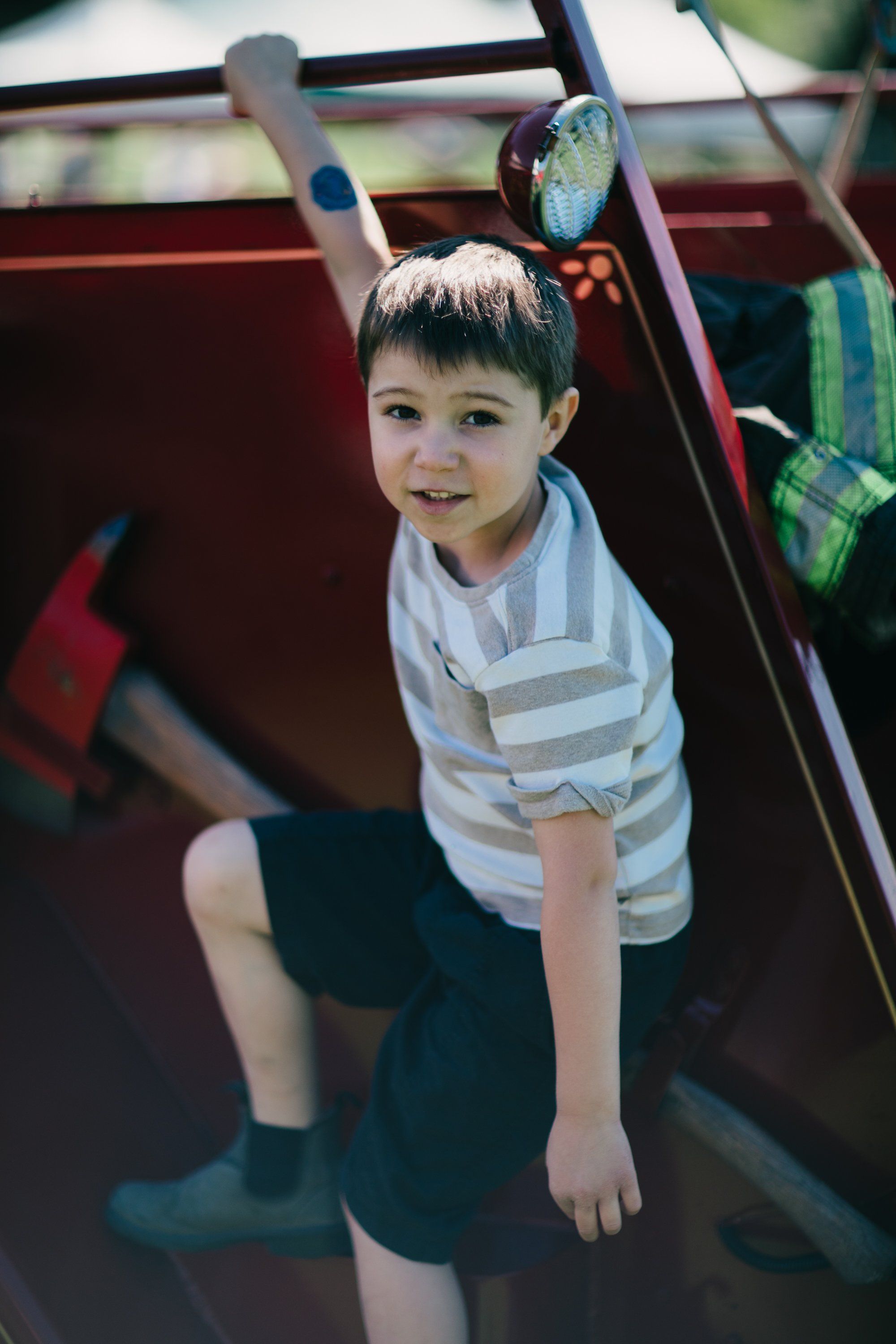 Young boy in striped shirt on wooden farm equipment, smiling at camera in dappled sunlight