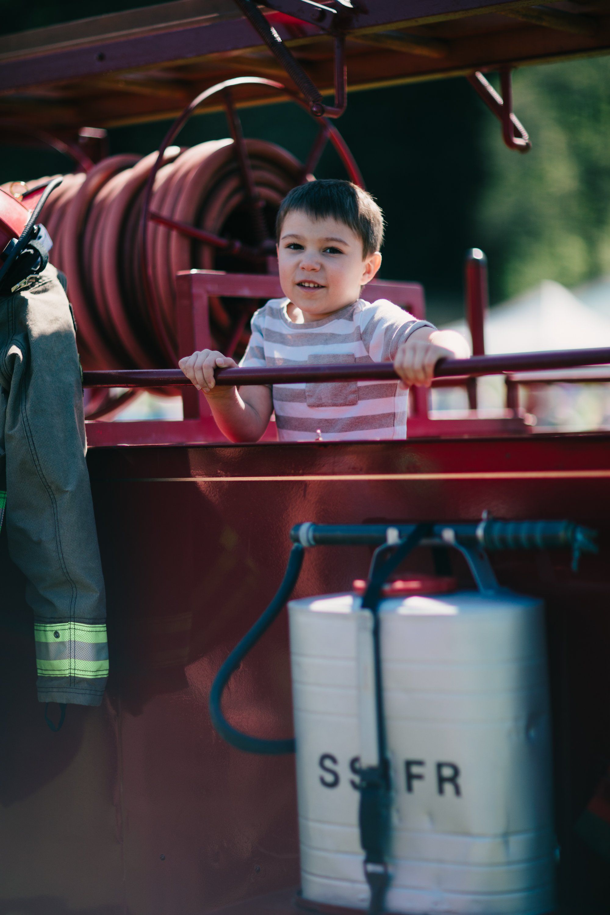 Toddler in striped shirt grips wooden farm equipment railing, smiling in warm sunlight with vintage machinery and pastoral blur behind