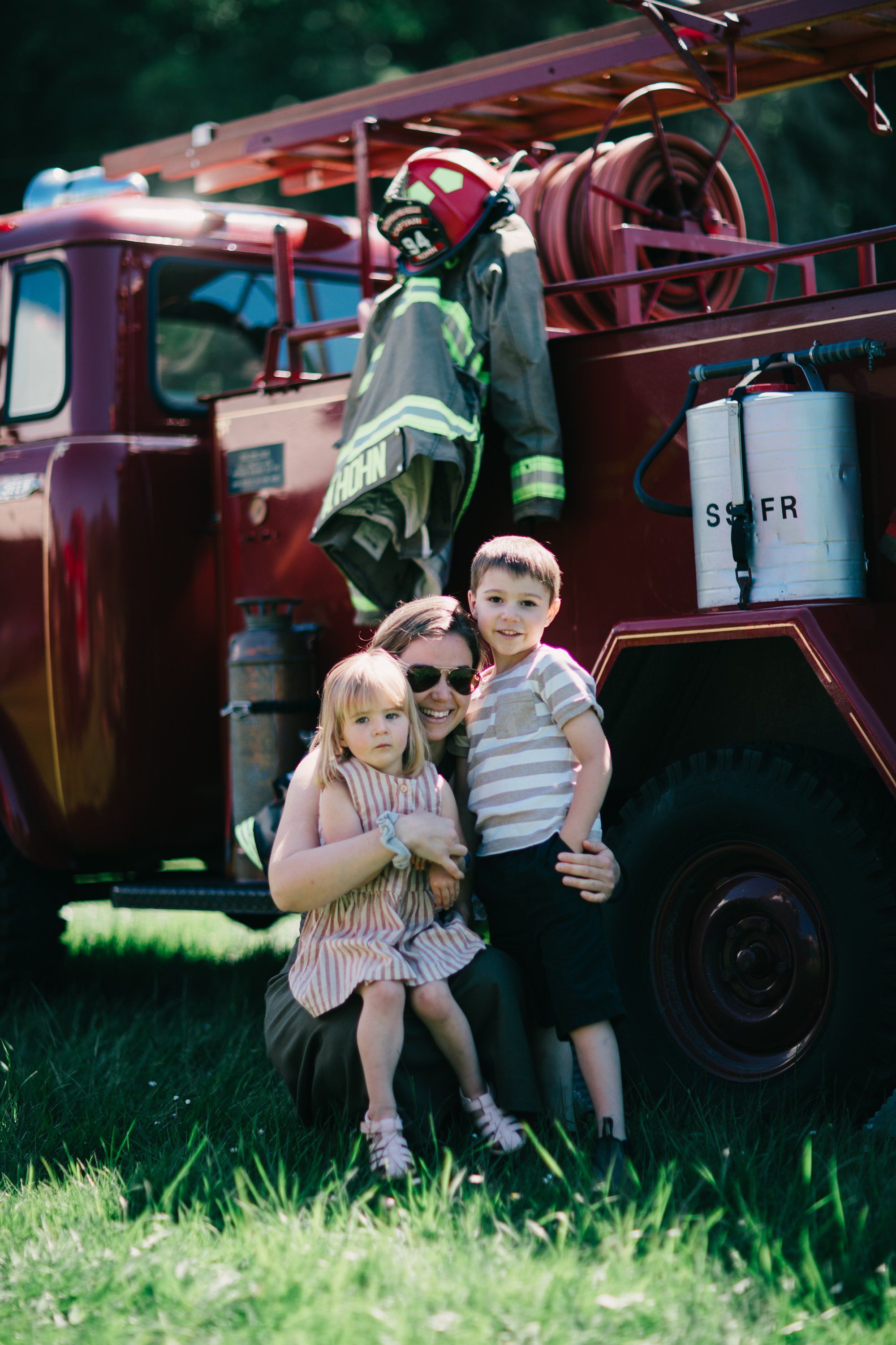 Two young children stand on grass beside a vintage red fire truck on a sunny day at Ruckle Farm