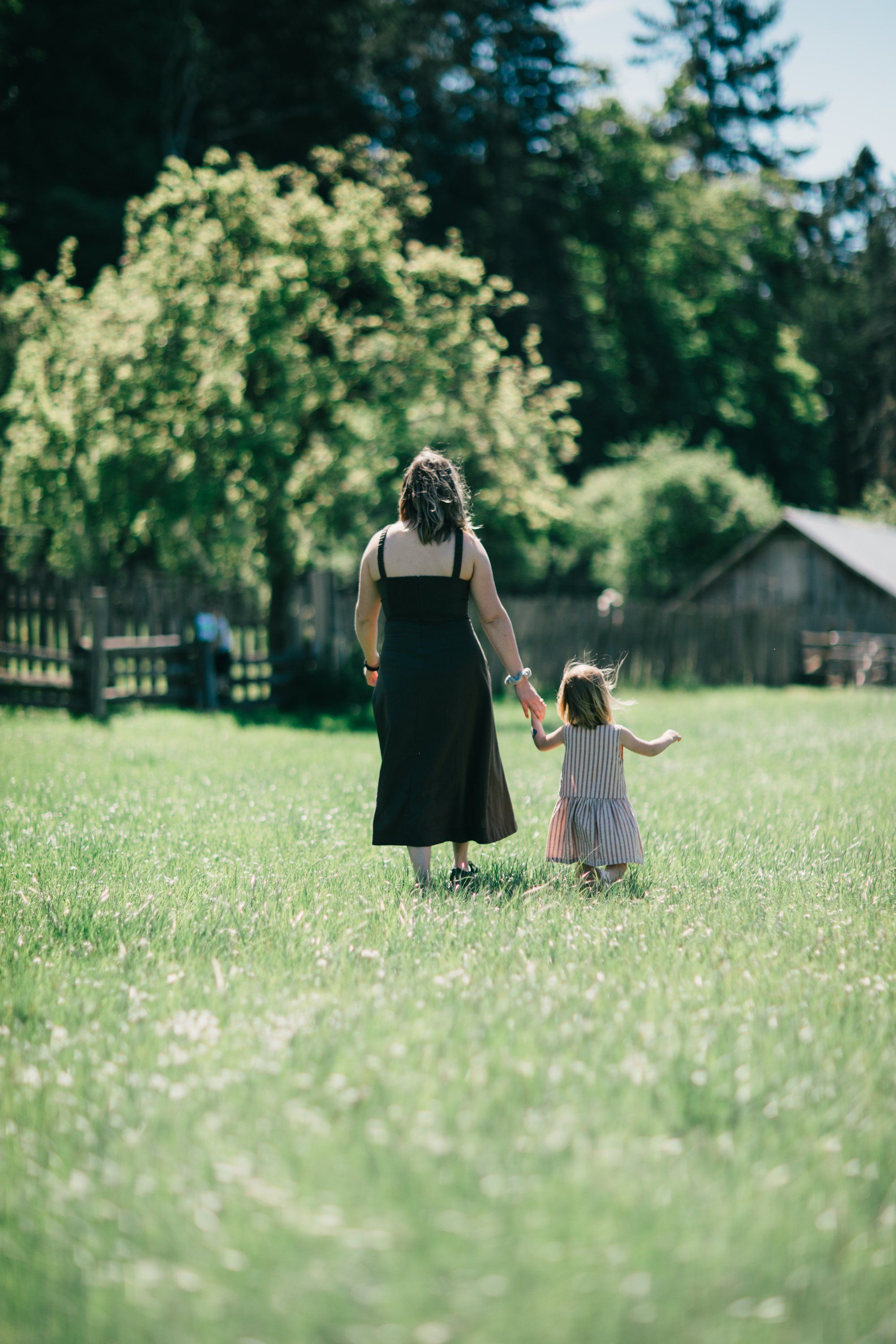Woman and young child hold hands walking through a sunlit meadow with wildflowers, framed by lush green trees and a distant barn
