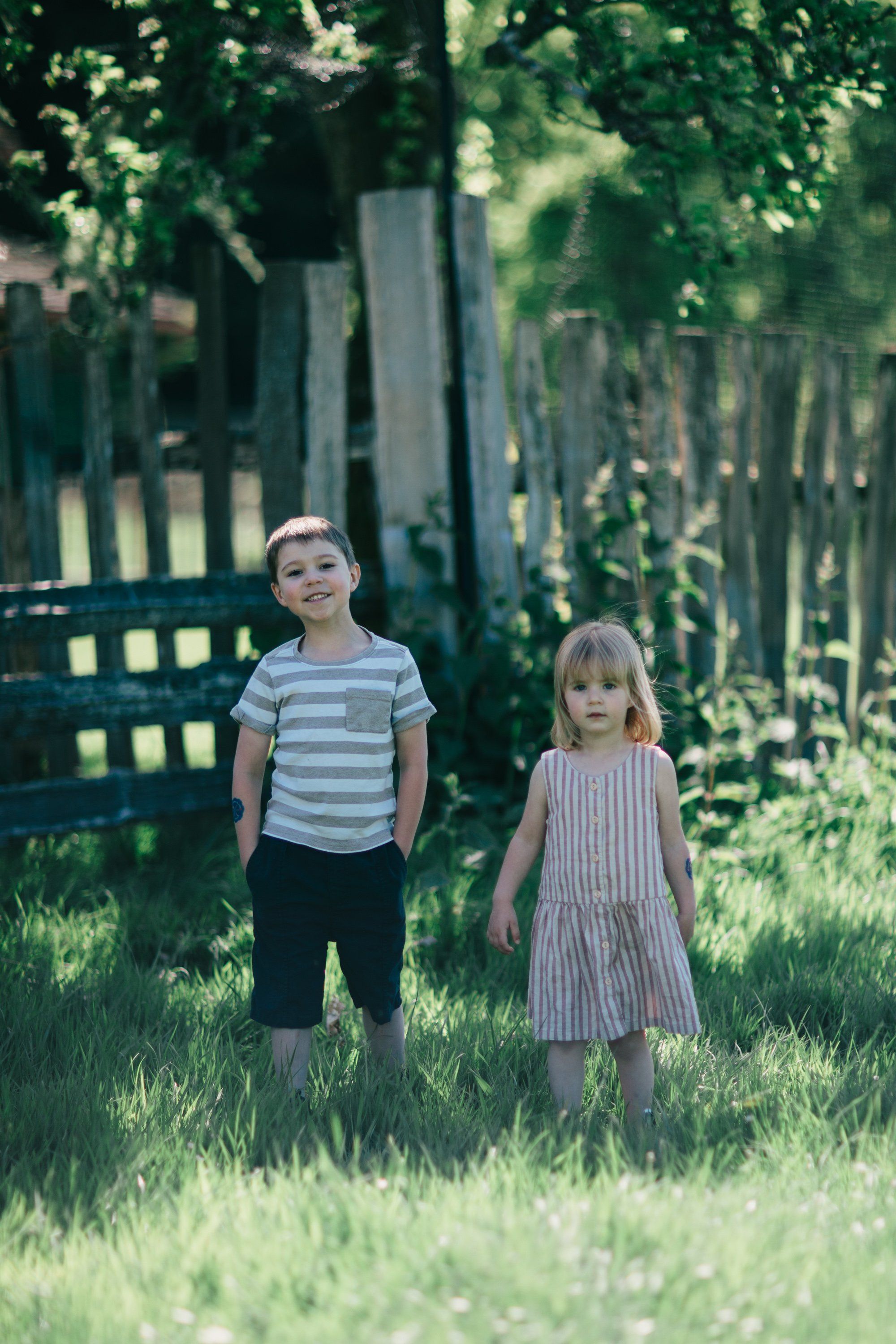 Two young children stand in tall grass beneath leafy trees, one wearing a striped shirt, the other in a pink dress, with a weathered fence behind them