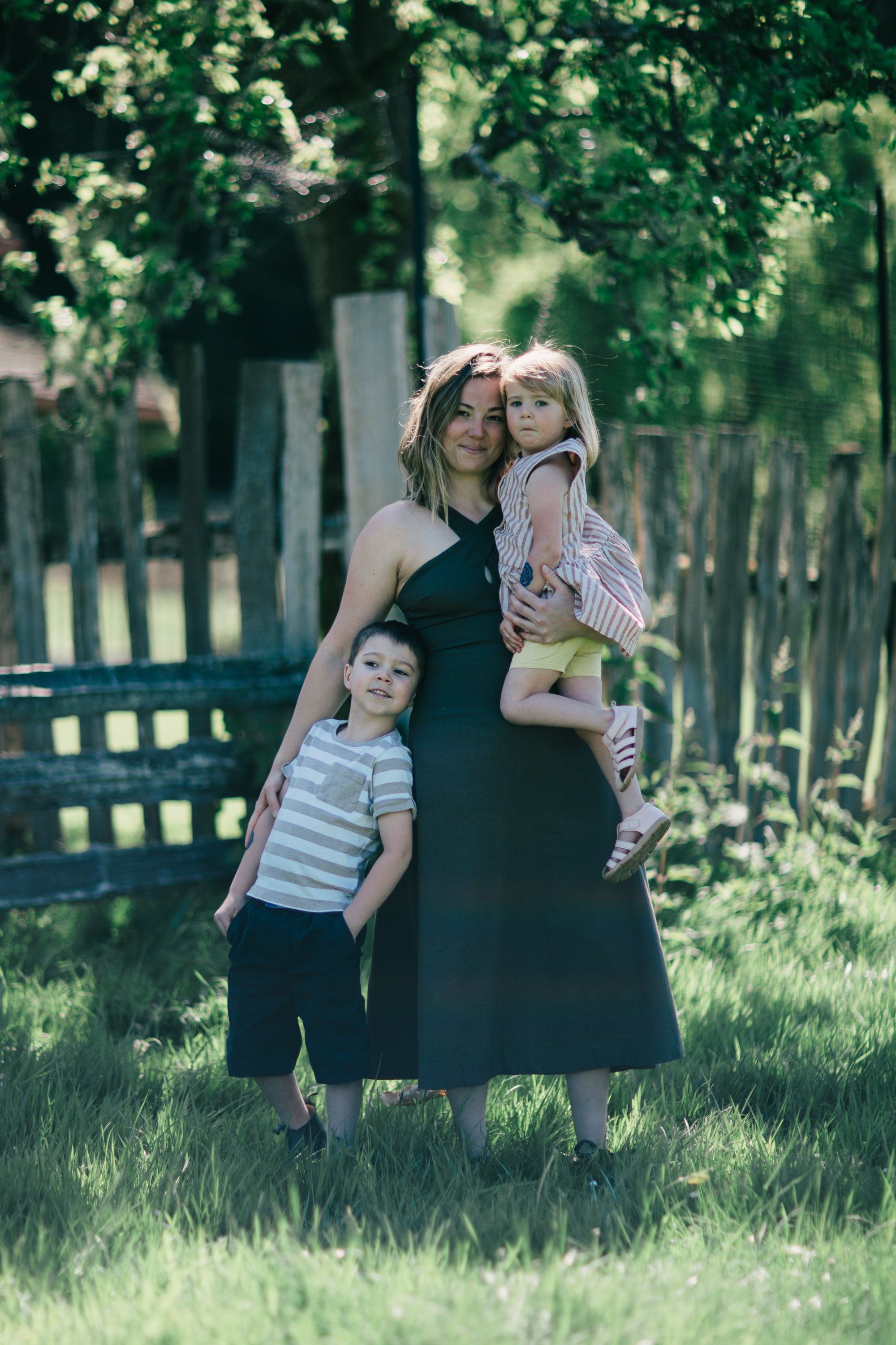 Woman holding two young children in sunlit meadow with white picket fence and tree canopy behind
