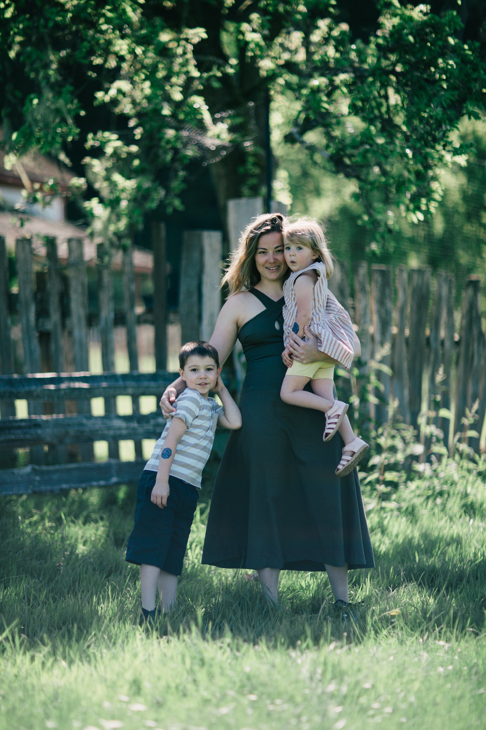 Mother with two young children standing in sunlit meadow beneath leafy trees and wooden fence