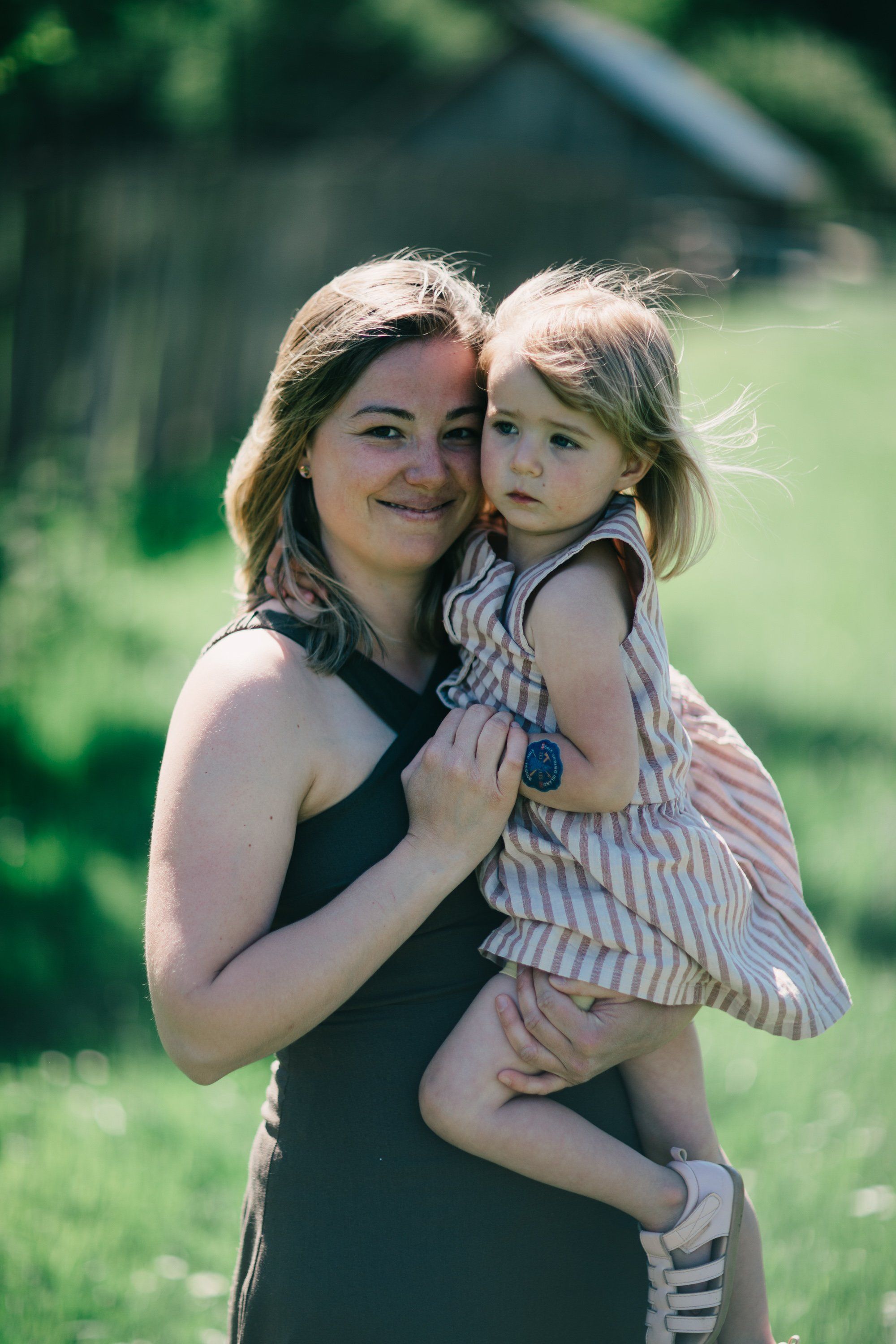 Woman holding toddler in striped dress in sunlit farmland, both smiling at camera with green fields behind