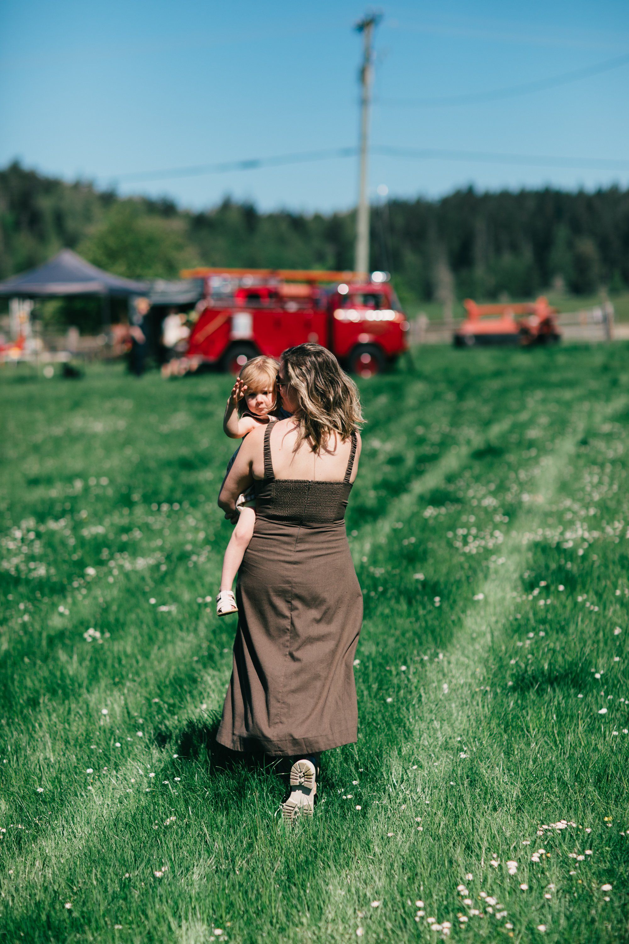 Woman holding toddler walking through wildflower meadow toward red farm equipment under blue sky