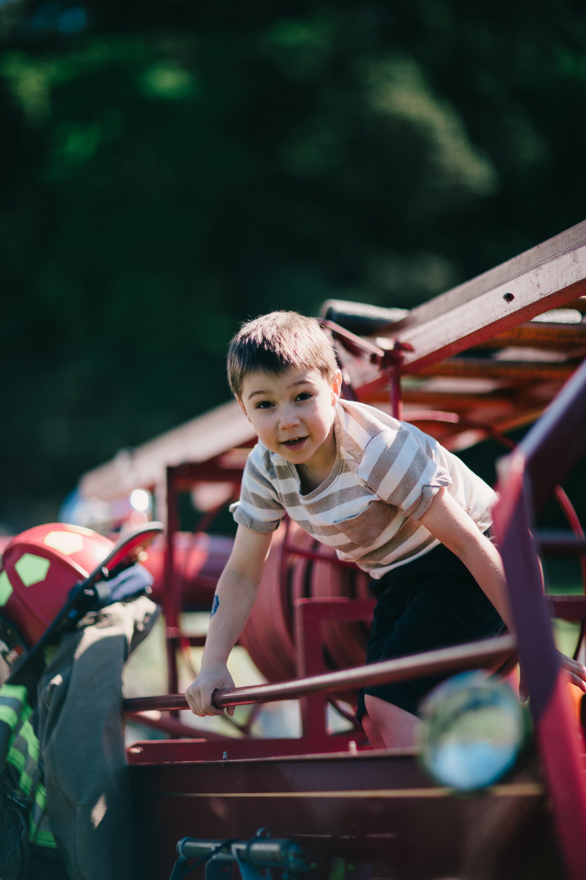 Young boy in striped shirt climbing on wooden farm equipment, sunlit meadow in soft focus behind him