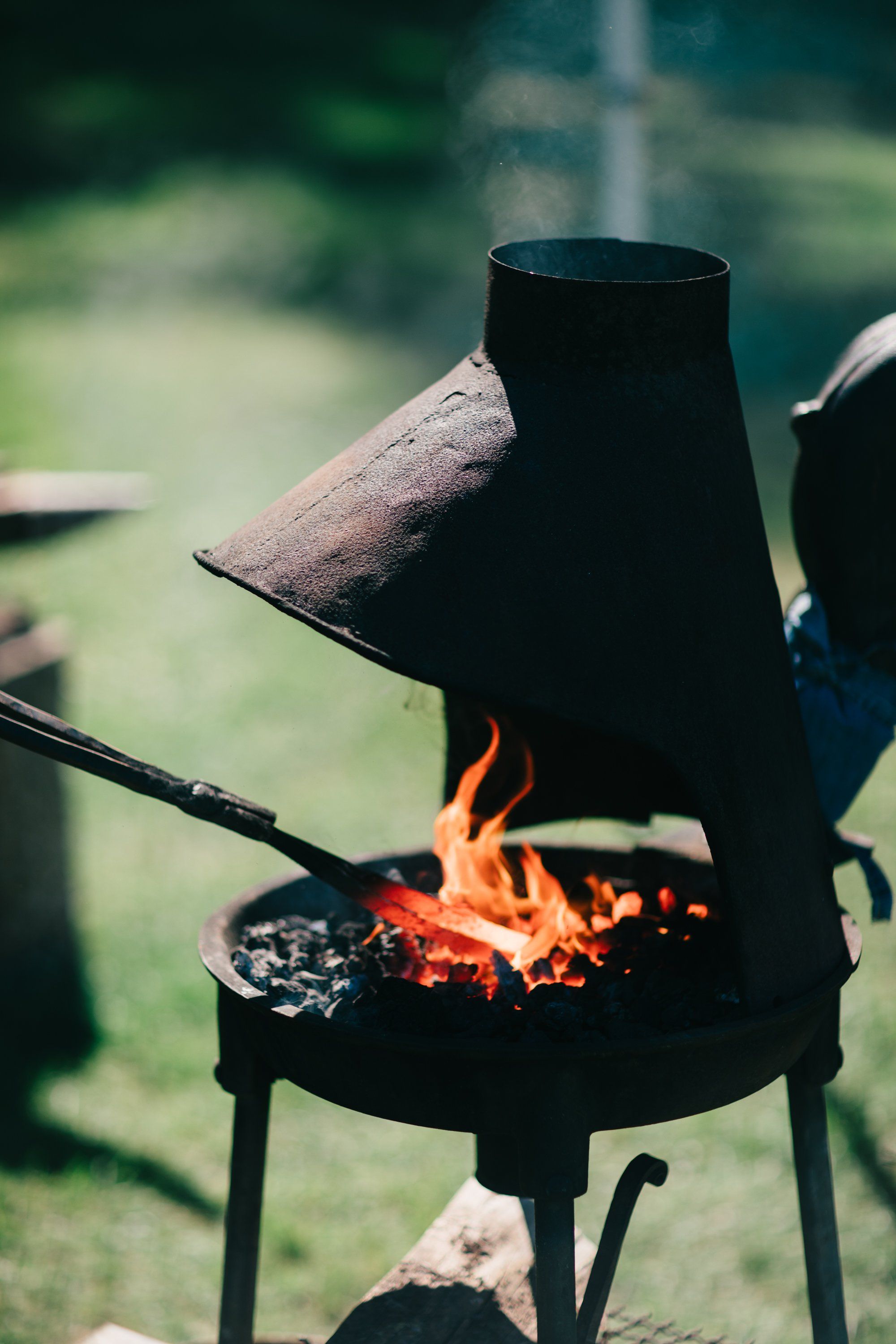 Outdoor wood-fired metal stove glowing with flames, a weathered log resting across the top, surrounded by soft green fields under natural daylight