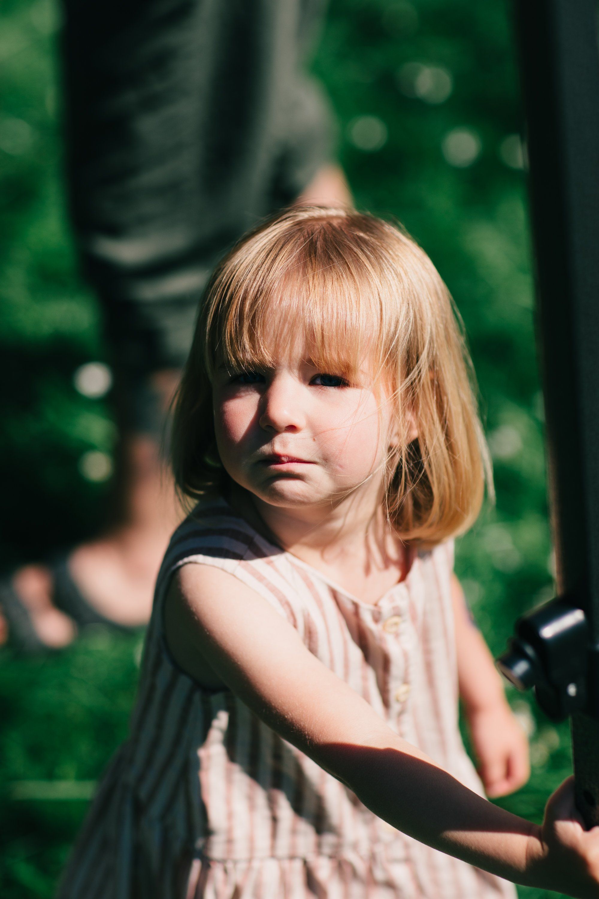 Young child with blonde bob haircut in pale dress gazes toward camera in dappled sunlight among green foliage