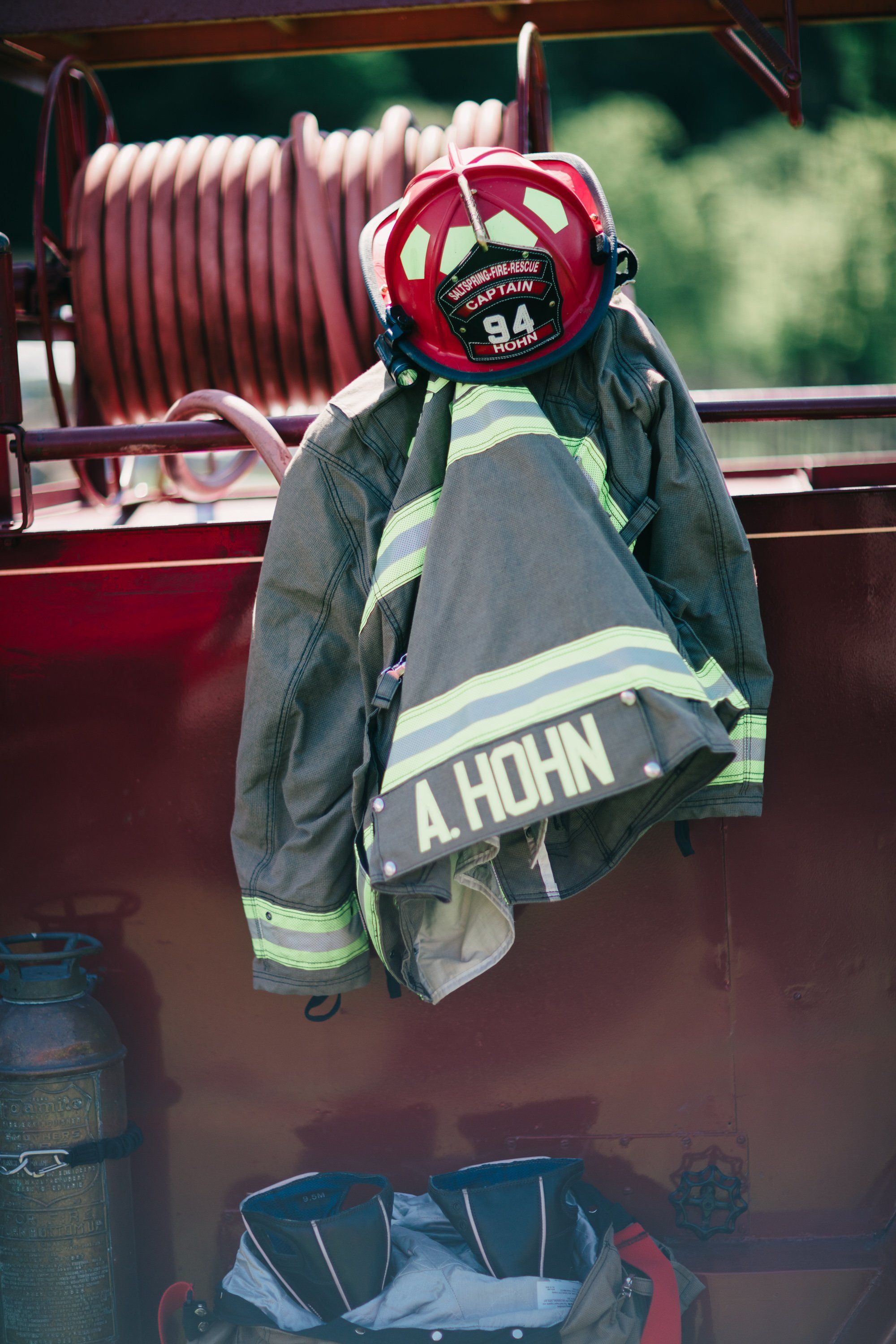 Firefighter gear hangs on a vintage fire truck, helmet and jacket with reflective stripes displayed against coiled red hose