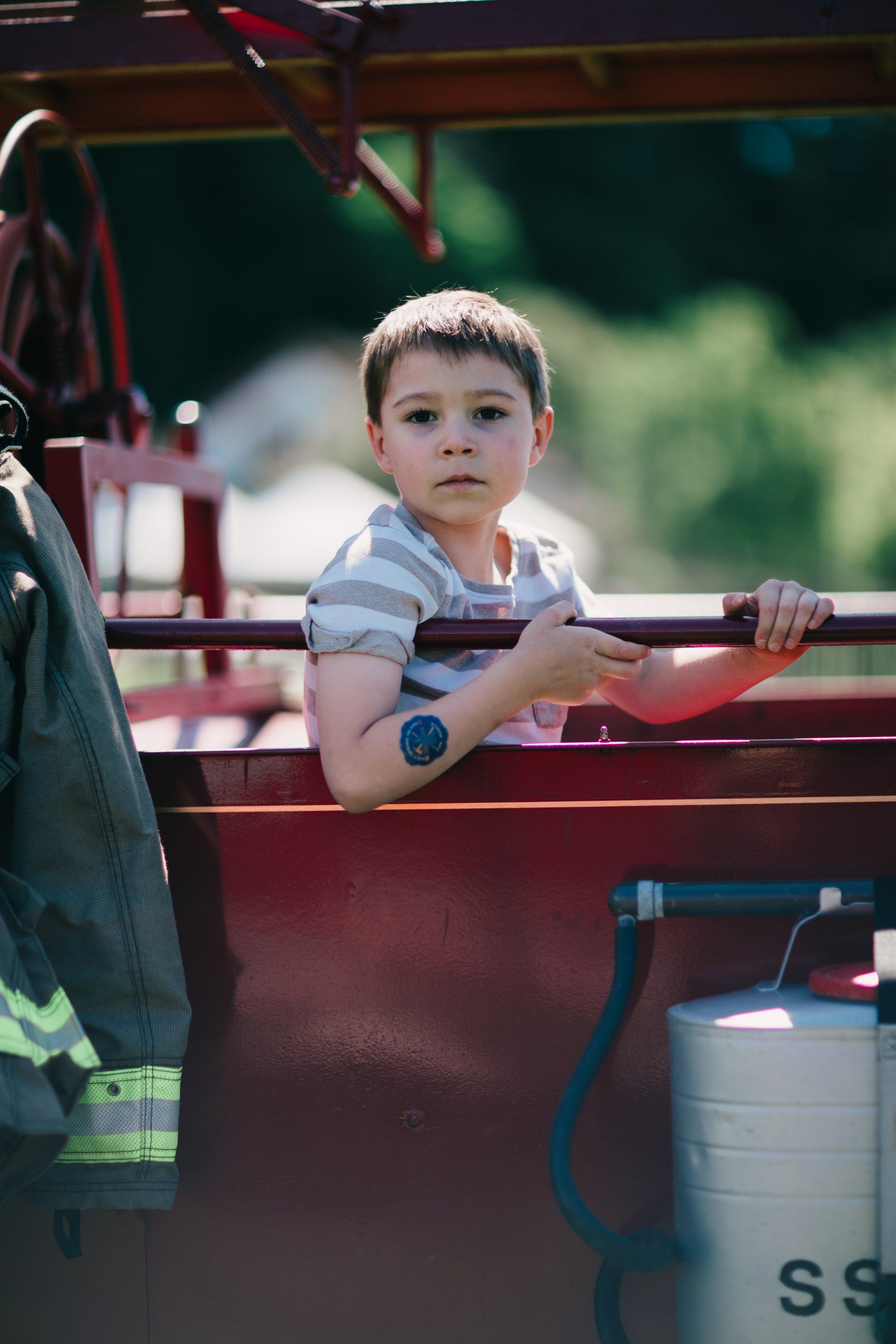 Young boy in striped shirt stands at wooden wagon rail, gazing thoughtfully amid farm equipment and dappled sunlight