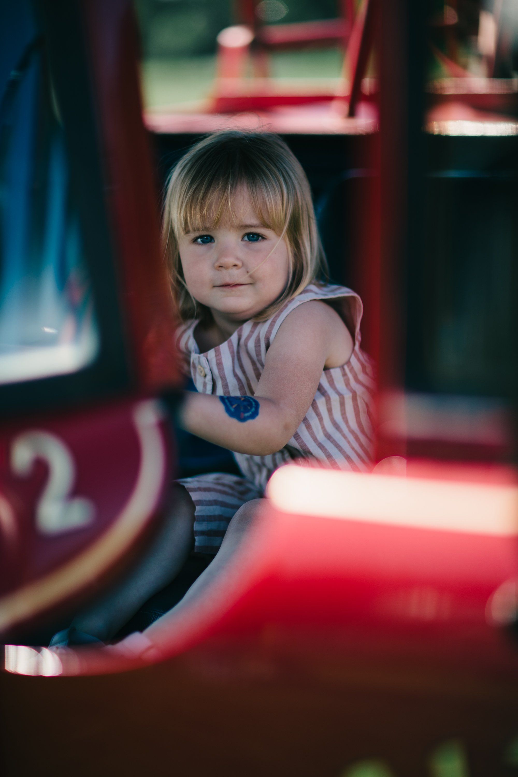 Blonde toddler in striped shirt gazes at camera among red carnival ride structures bathed in warm neon light