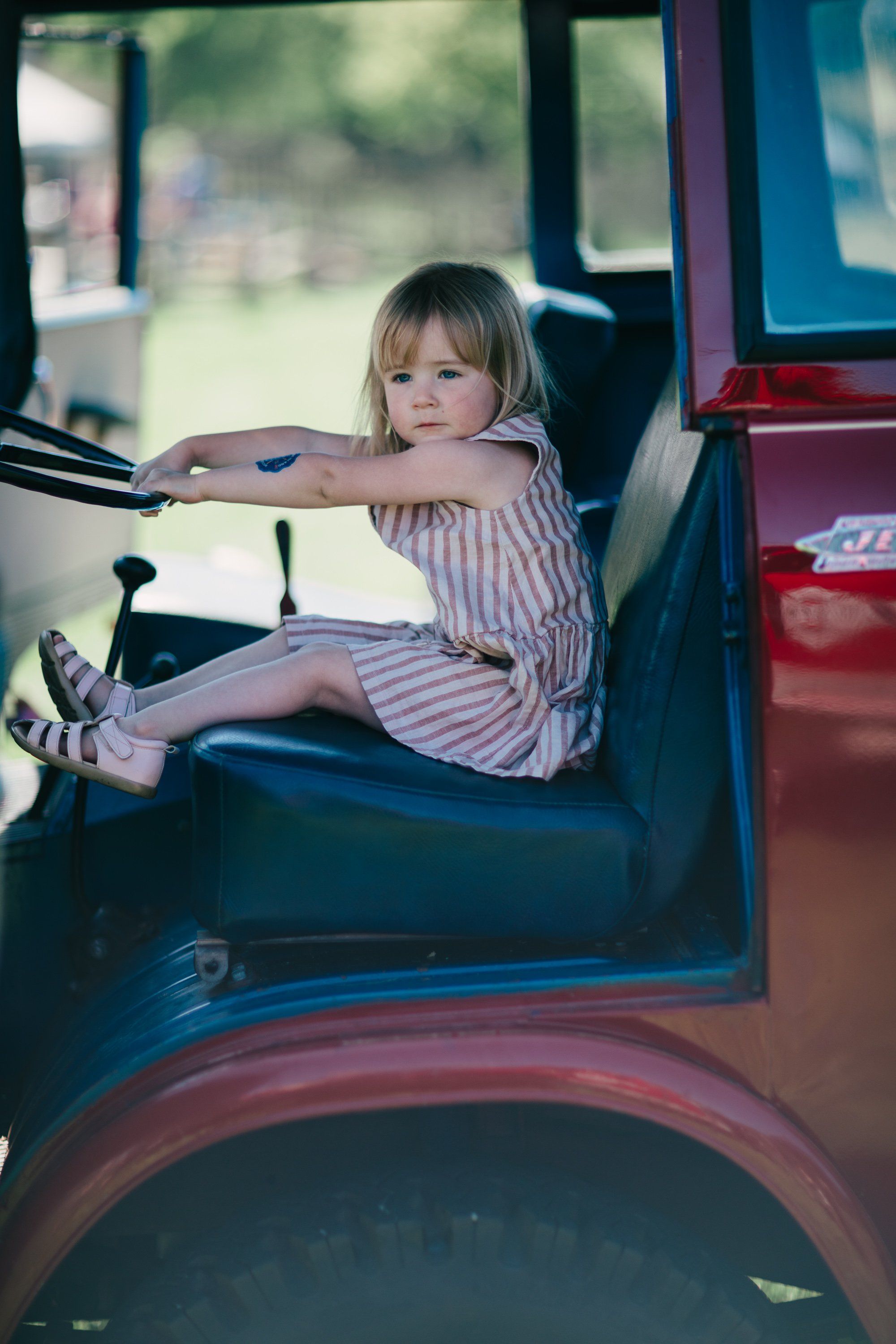 Young child at the steering wheel of a vintage truck, hands extended, wearing striped shirt and sneakers, warm afternoon light