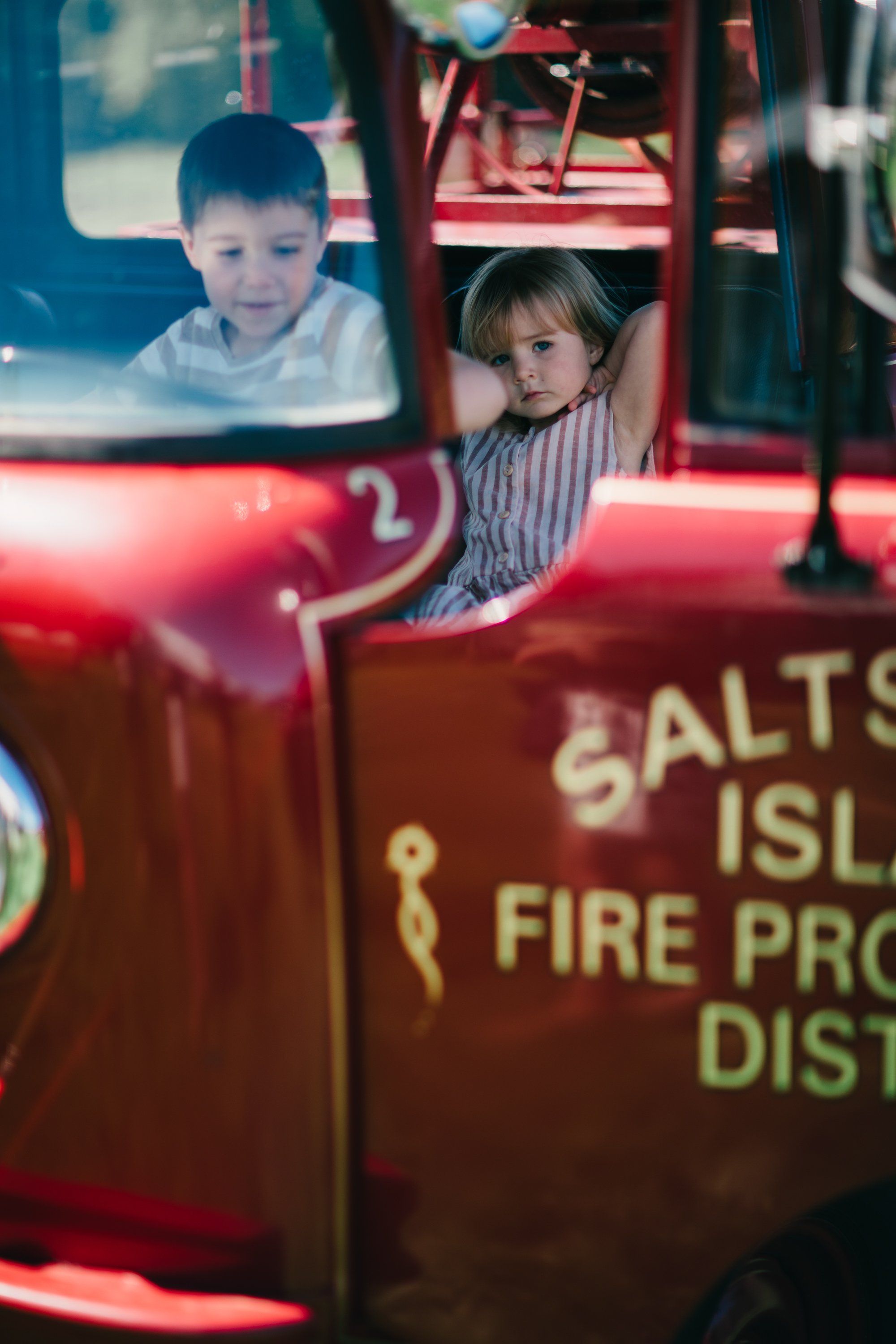 Two children peer from the windows of a red fire truck, sunlight glinting off the glass at a farm event
