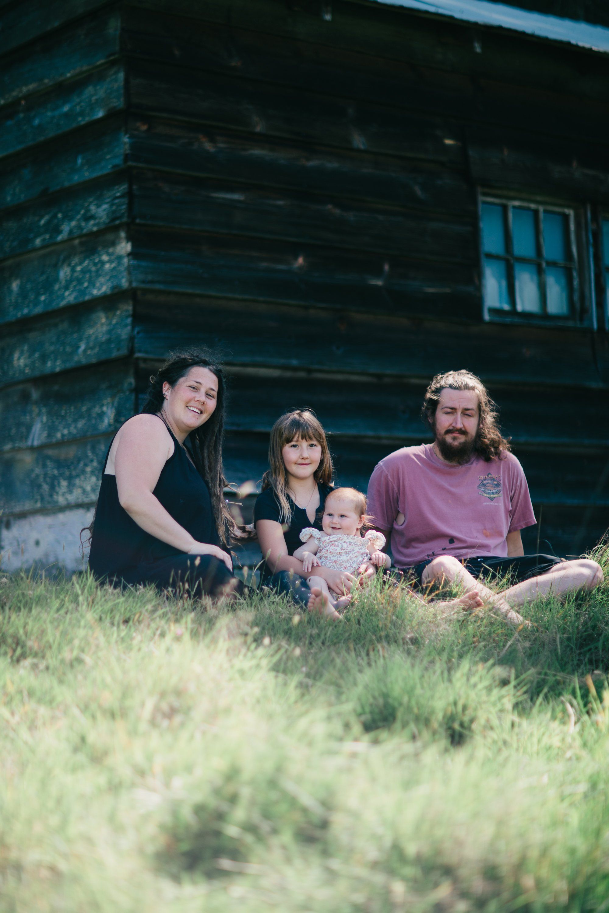 Family of four sitting in tall grass beside a weathered log cabin with blue-trimmed windows, soft natural light casting a warm, intimate mood