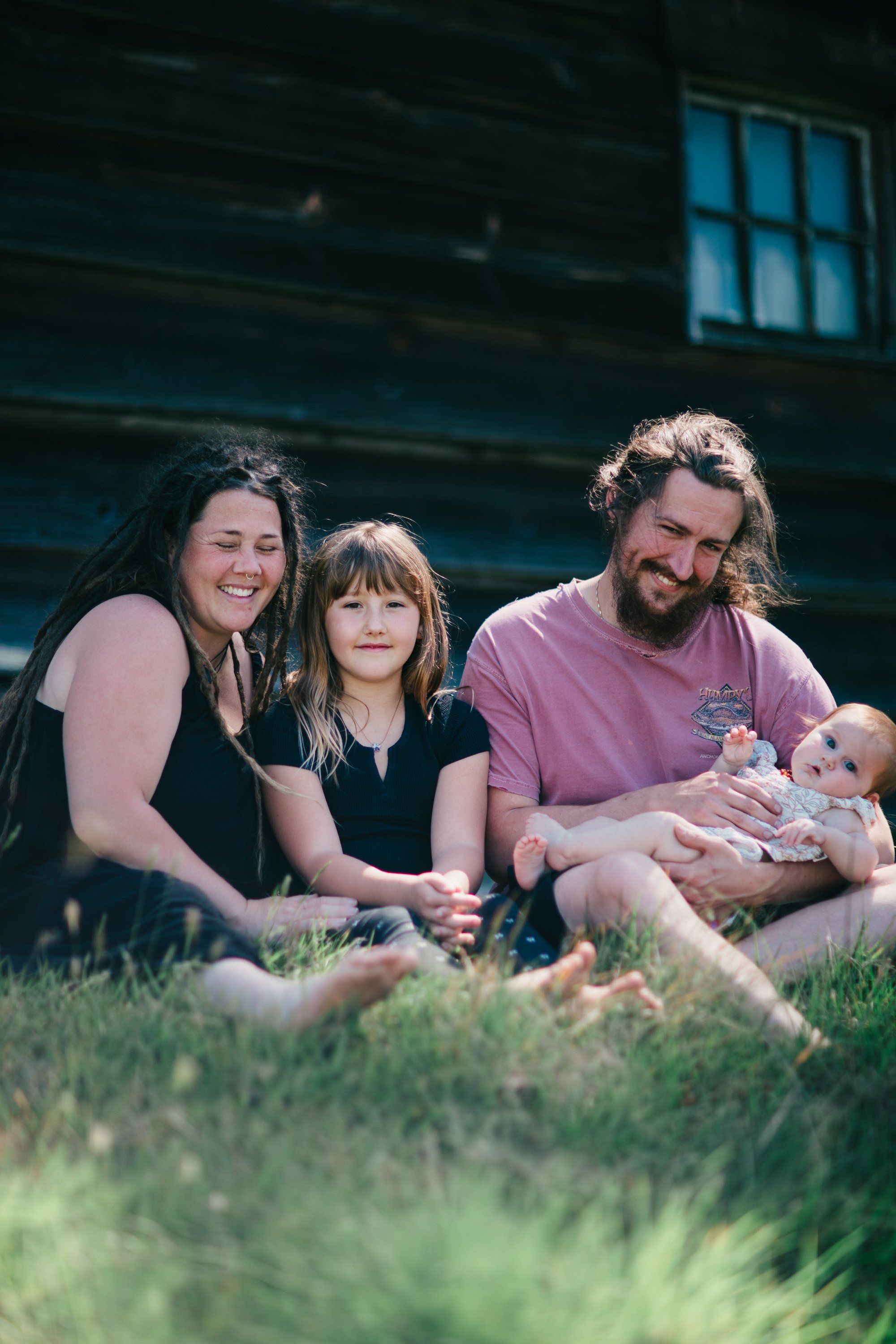 Family of four sitting on grass at Ruckle Farm with wooden barn and blue window behind them in golden evening light
