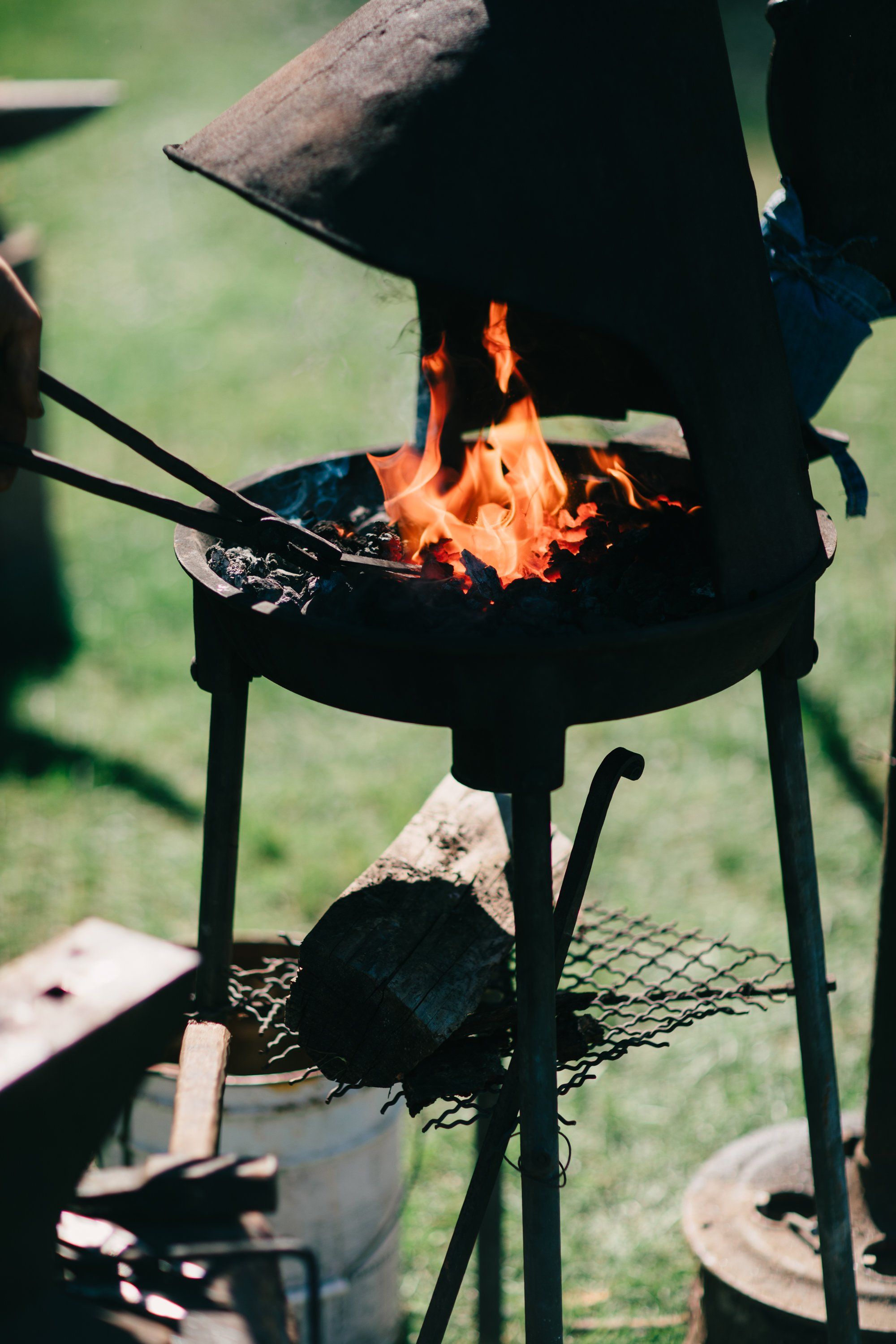 Flames leap from a black wood-fired cauldron suspended over glowing coals, with a log resting across the top and green grass visible beyond