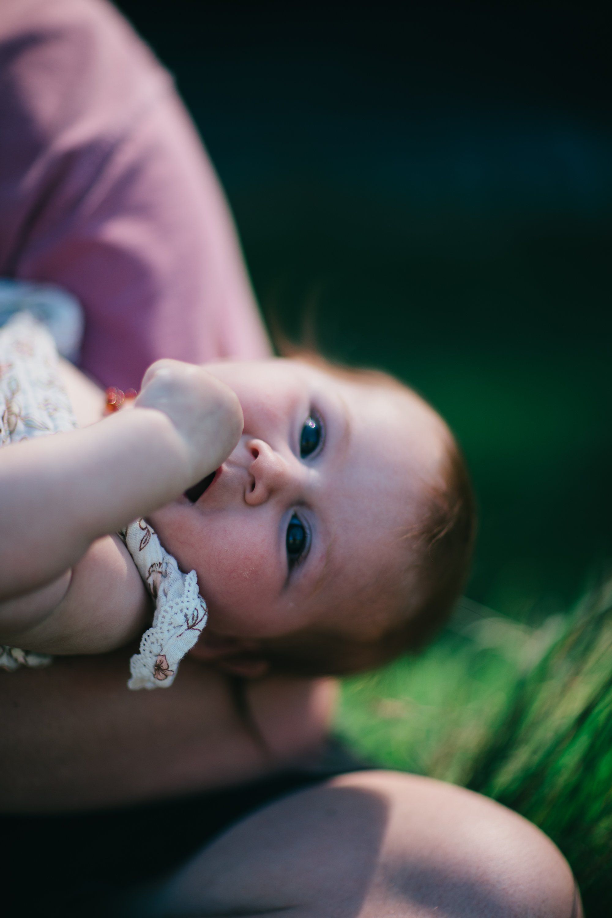 Newborn baby in white lace lying on purple fabric gazes upward, soft natural light and blurred green field background