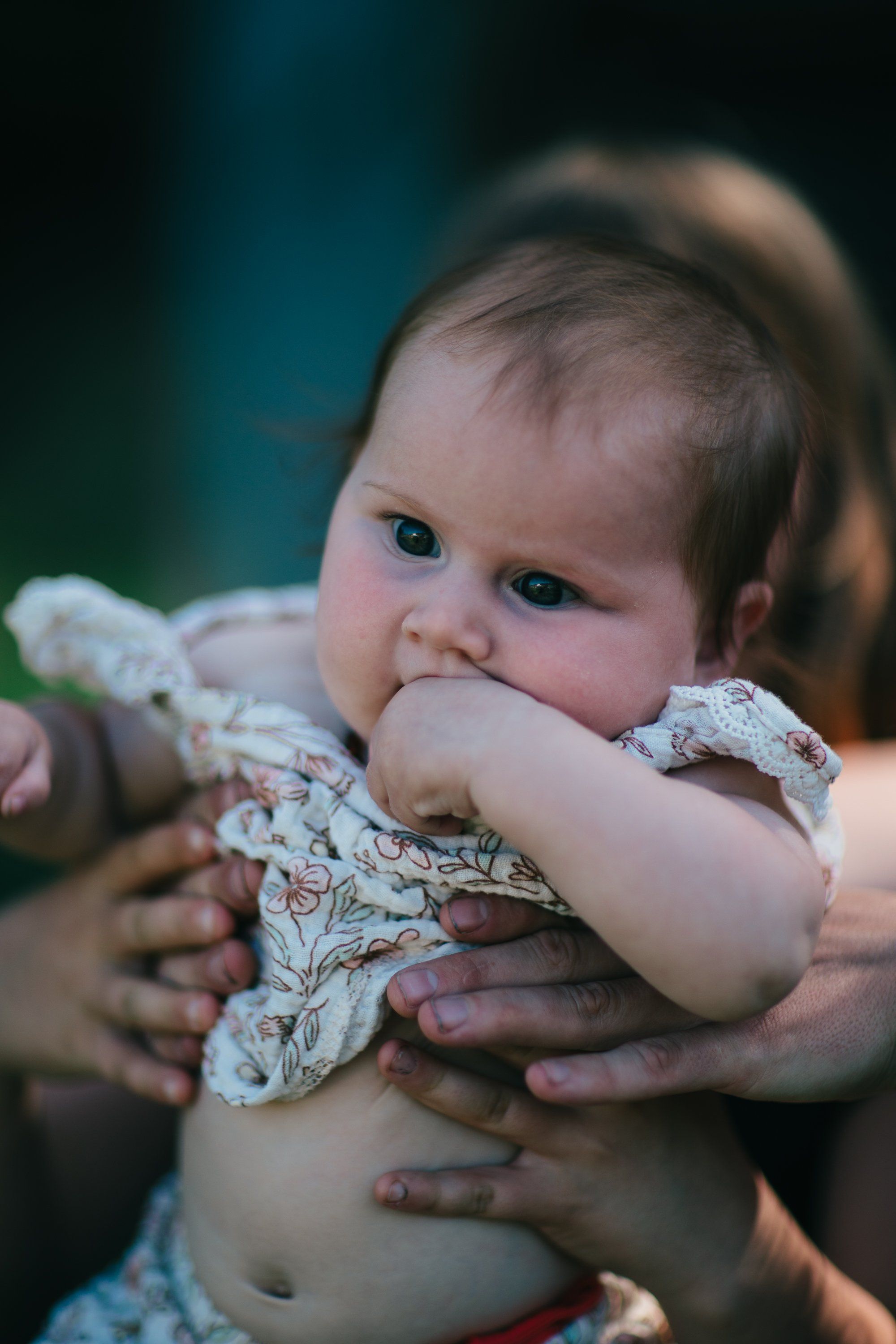 Toddler in patterned shirt chewing hand, held by caregiver against soft-focus green background