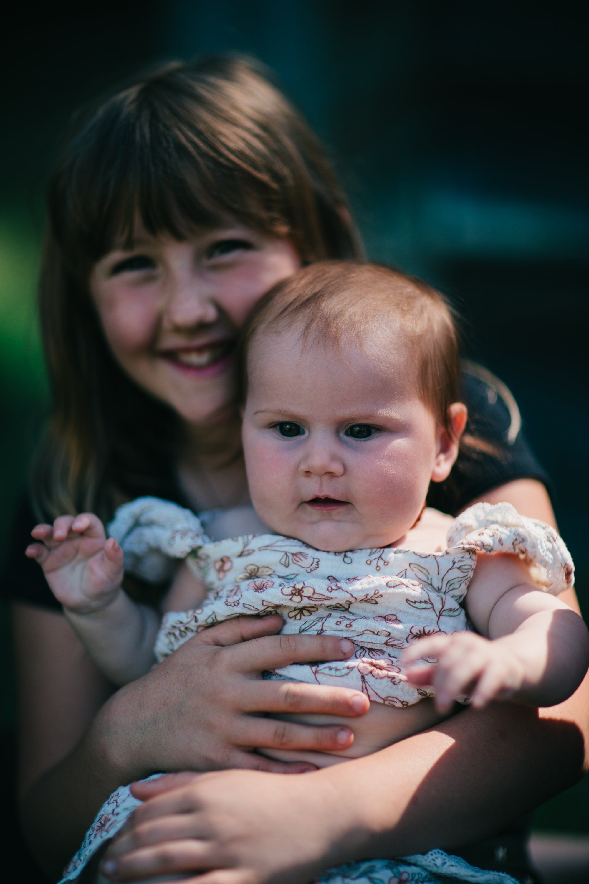 Older girl smiling at camera while holding a toddler in a patterned dress, warm sunlight illuminating their faces against a soft, blurred teal background
