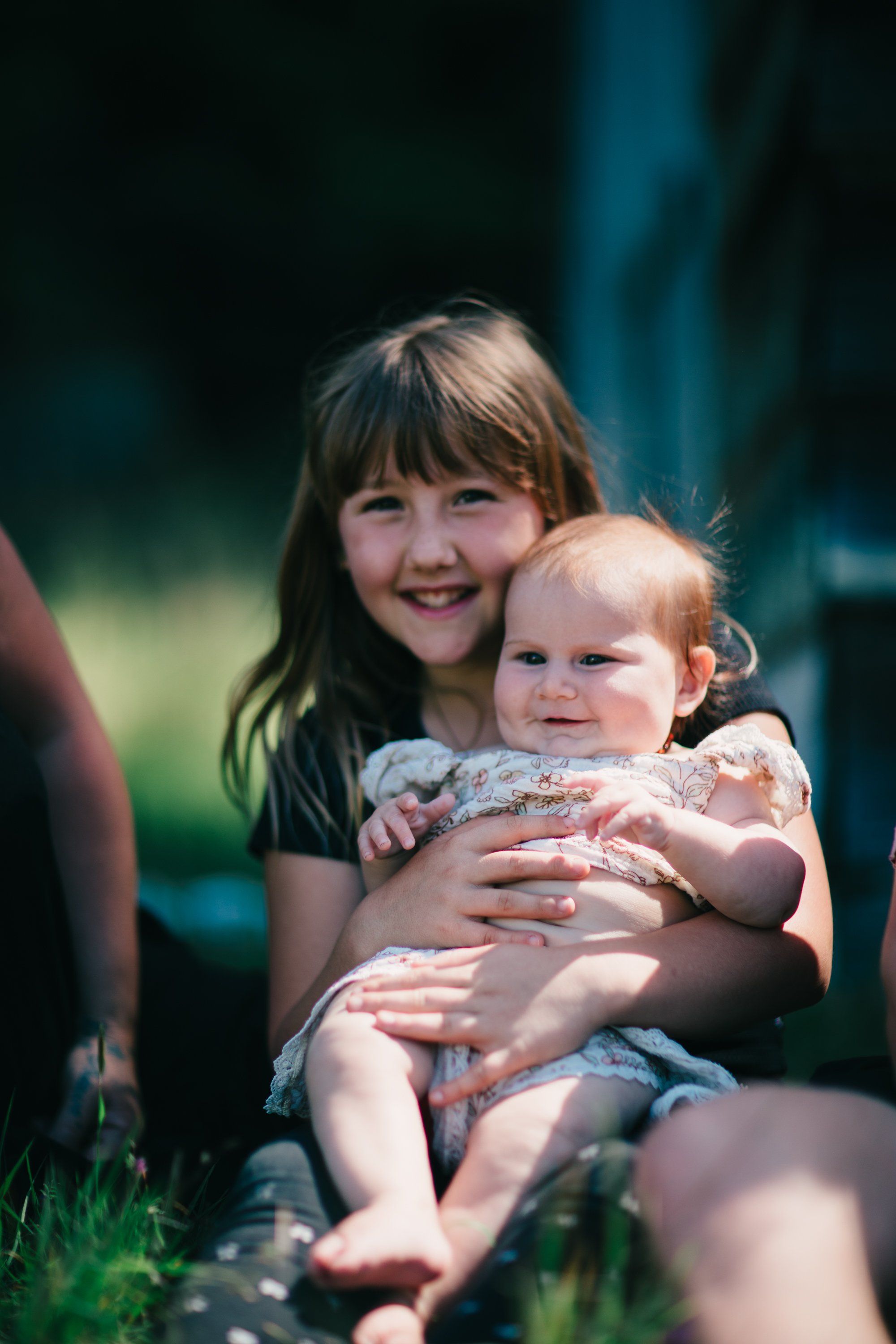 Older girl holding smiling toddler outdoors, dappled sunlight on grass, warm family moment at farm setting