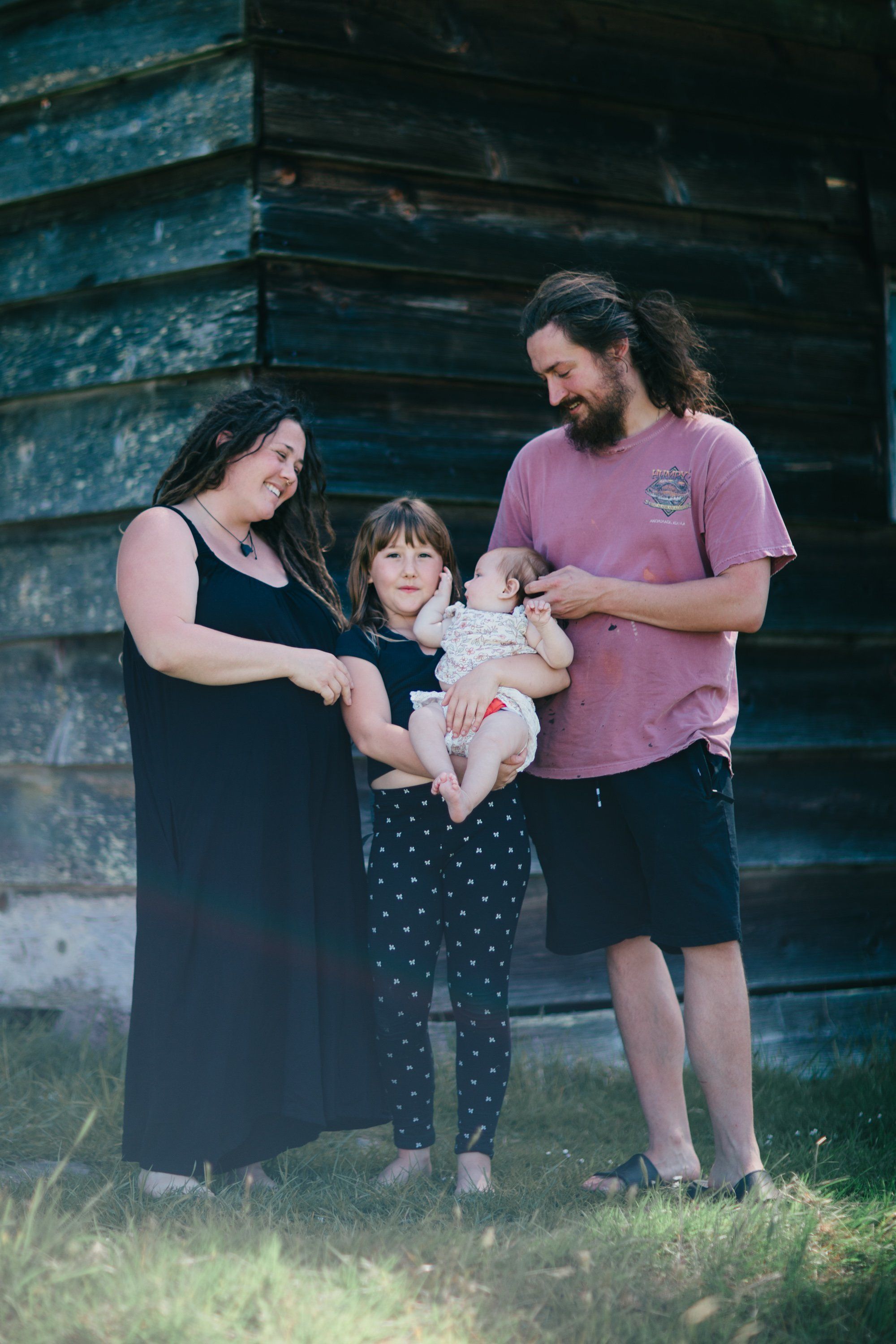 Family of four holding a baby in front of weathered blue wooden farm building on golden afternoon light